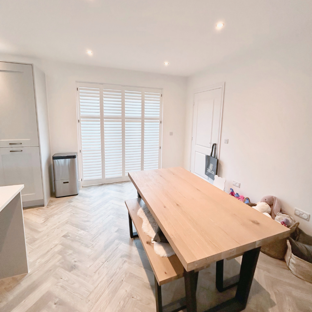 Dining area with a wooden table, bench, and chairs, white walls, window with shutters, and some toys in the corner.