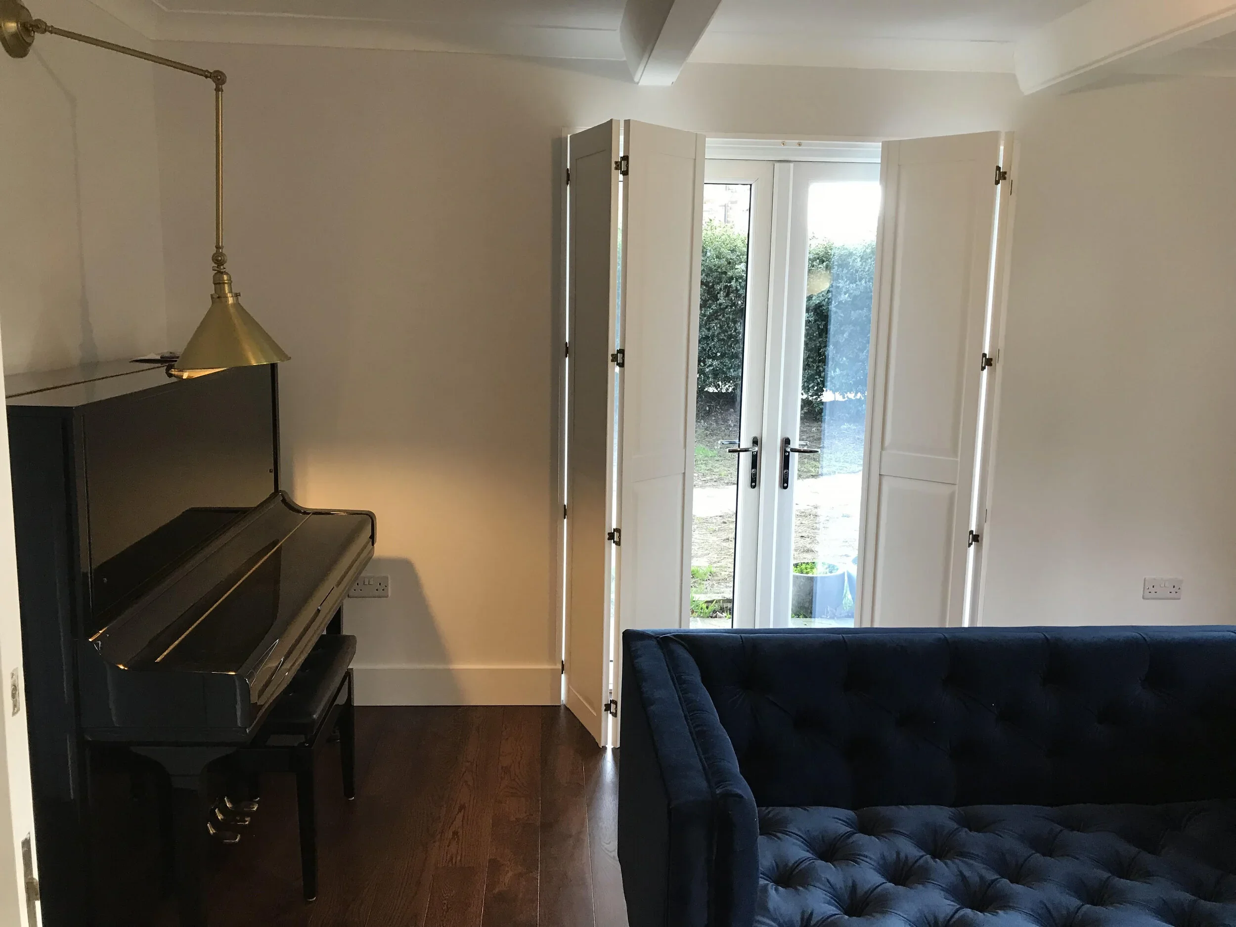 A room with a solid panel shutters, a black upright piano, a gold-colored lamp, and a dark blue tufted sofa. Open shutters reveal greenery outside.