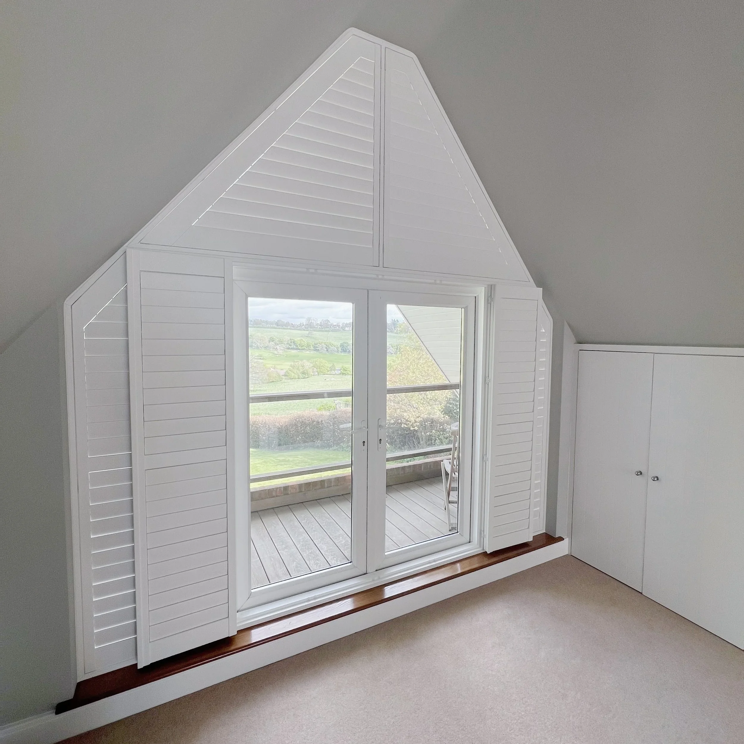 Interior of a room with a large window and white wooden shutters in an attic space with a sloped ceiling.