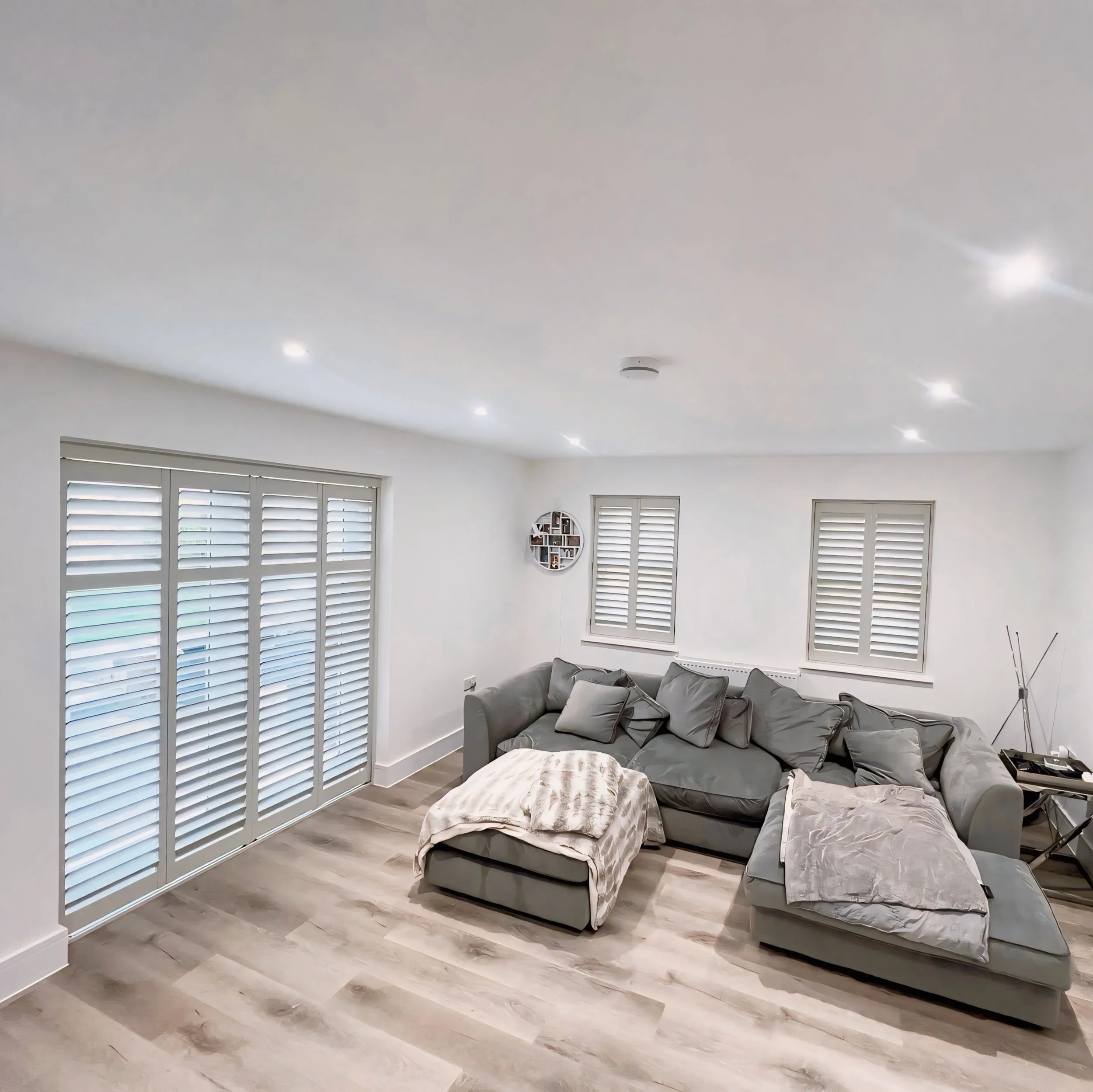 A living room with closed white shutters on windows, white walls, a gray sofa with gray pillows, and scattered plastic-wrapped furniture or objects on the floor. There are round wall shelves with small decorative items on the wall.