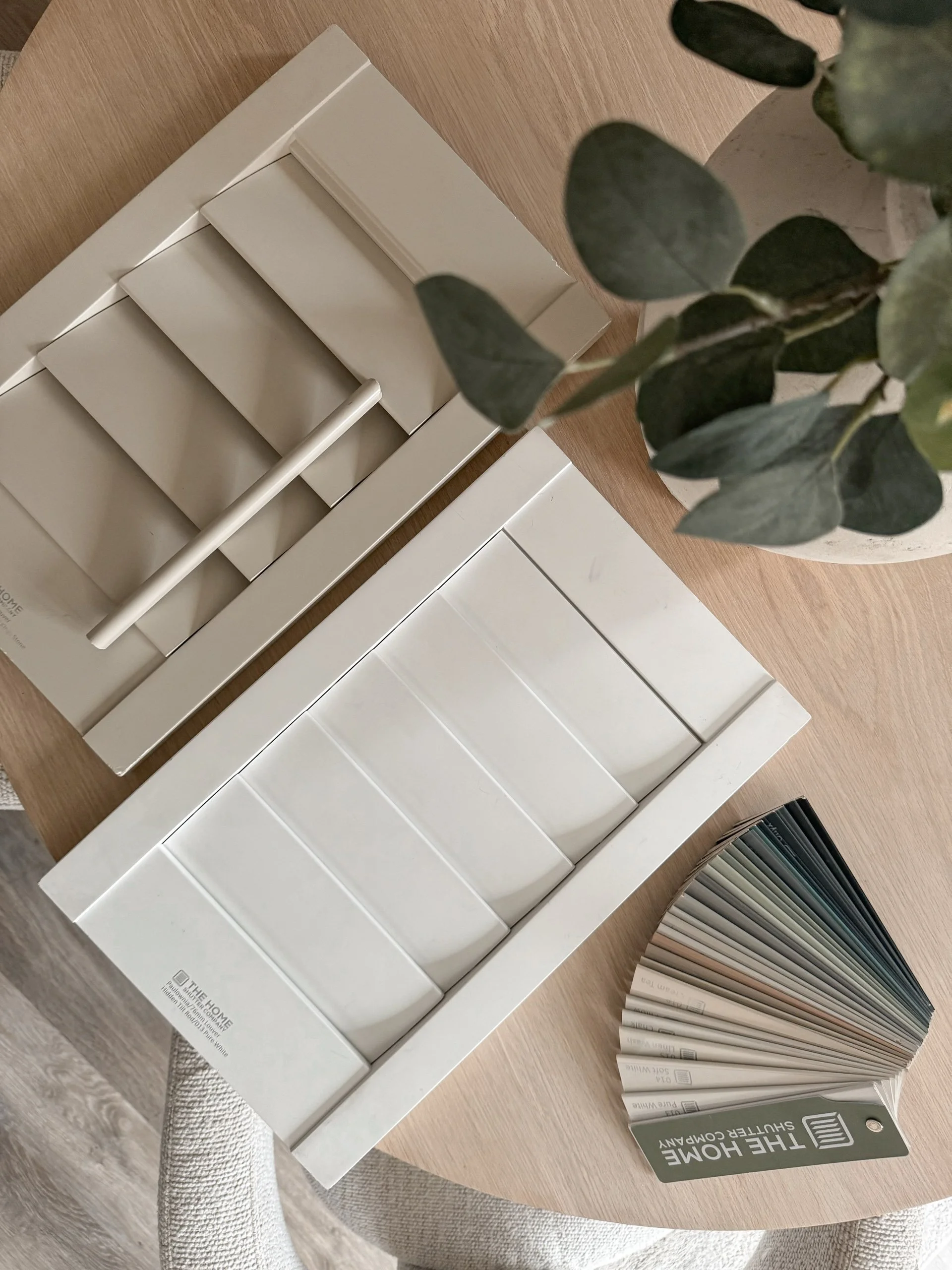 Swatch fan deck, color sample book, and paint sample tiles on a round wooden table. A white sample box and a small potted plant are also visible.