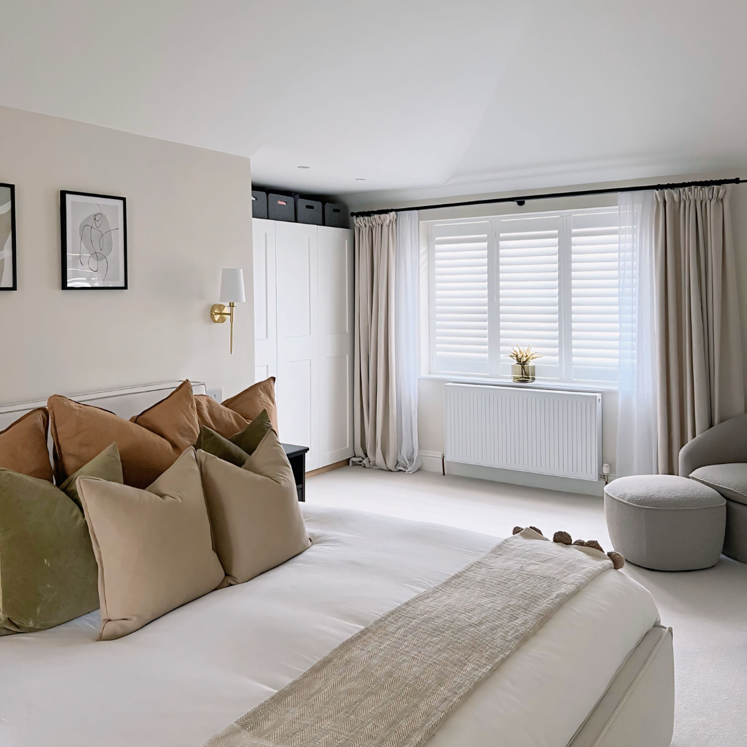 Bright bedroom with white walls, window with horizontal blinds and curtains, beige bed with multiple pillows, a small black side table, a gray armchair with an ottoman, and minimal wall decor.