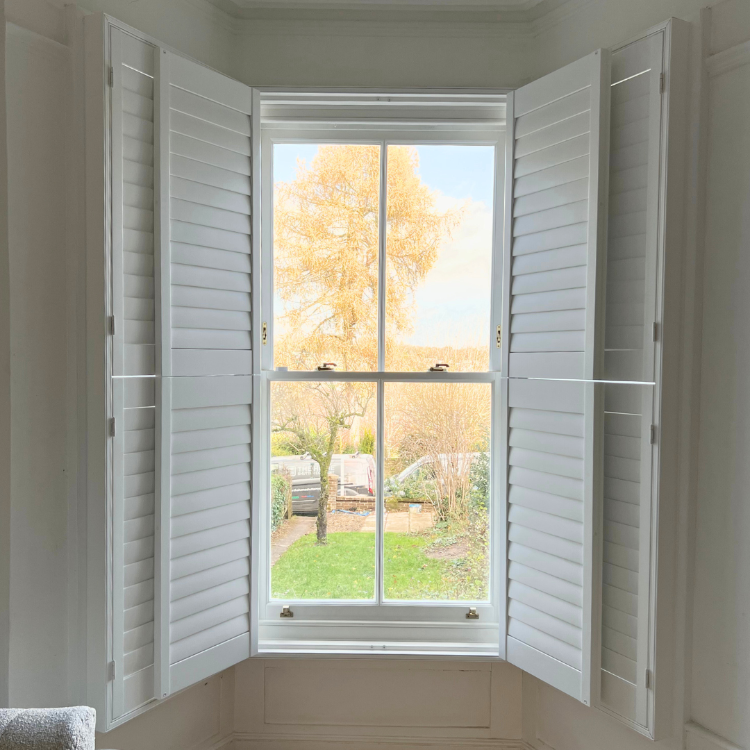 A view through a window with white shutters, showing a tree with orange leaves in a yard with grass and various plants.
