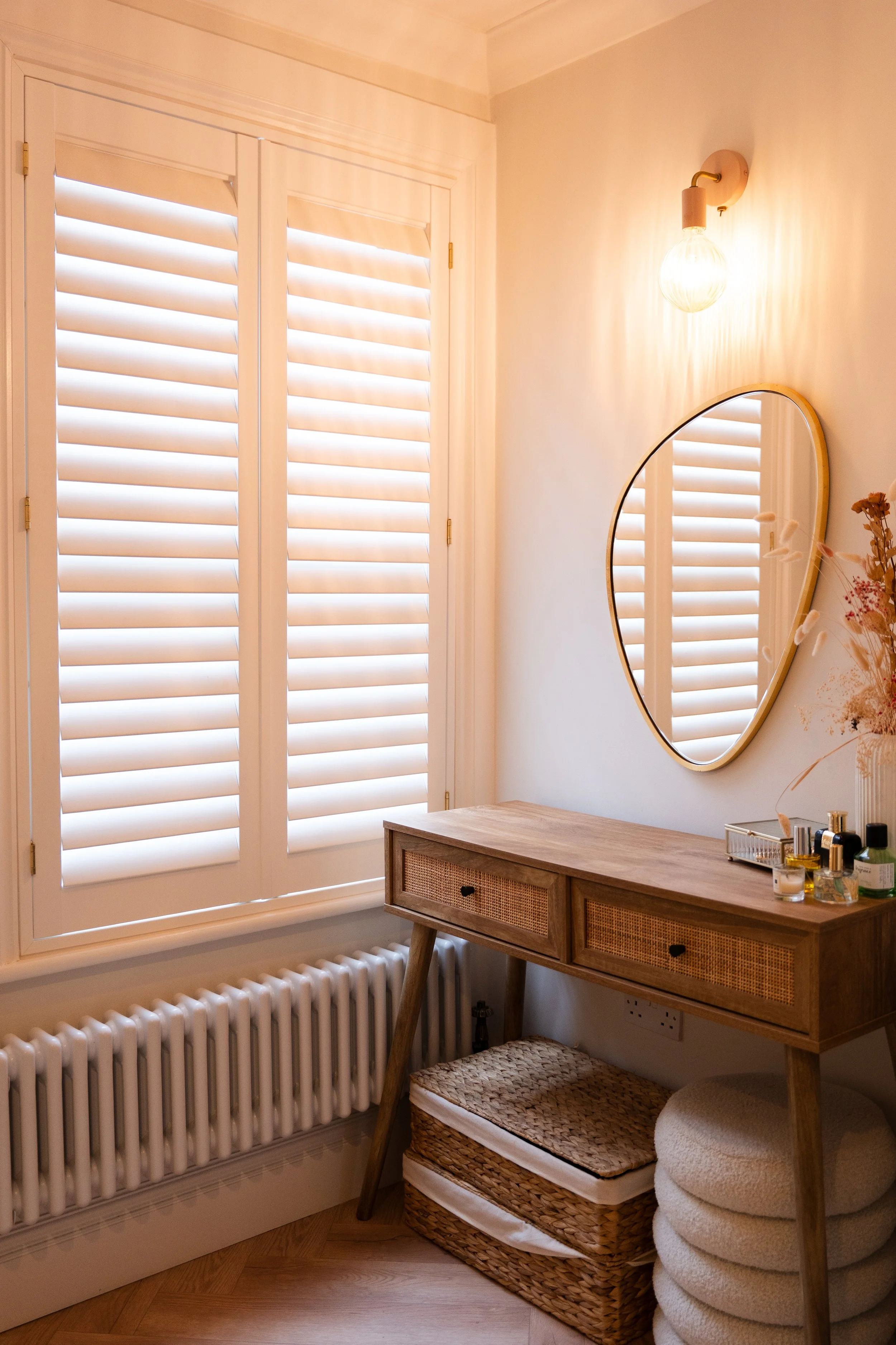 A cozy corner of a room with a window covered by white plantation shutters, a round mirror on the wall, a wooden desk with wicker drawers, and items like bottles and skincare products on the desk. There are baskets and padded stools beneath the desk,