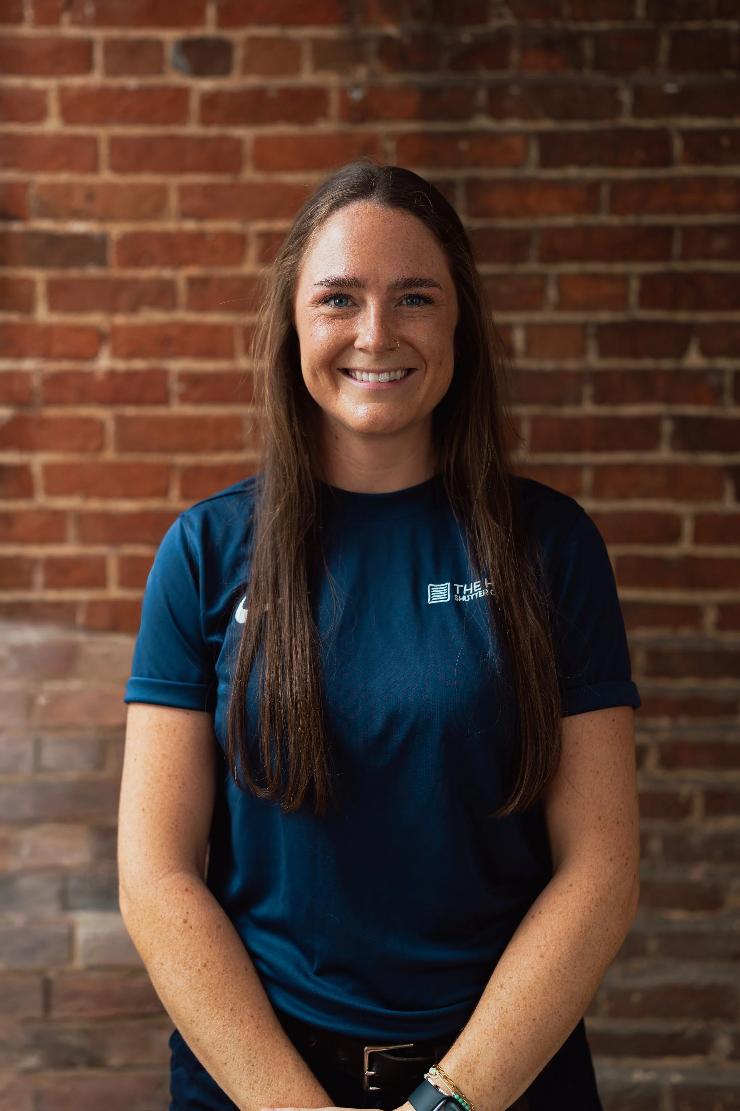 A woman with long brown hair, blue eyes, and a nose piercing smiling in front of a red brick wall. She is wearing a navy blue T-shirt with a logo and has a watch and bracelets on her left wrist.