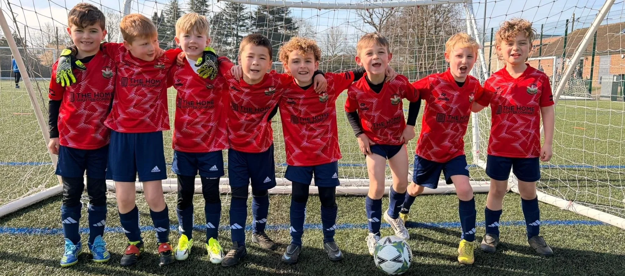 A group of young boys in red soccer jerseys and shorts standing in front of a soccer goal on a field, smiling with their arms around each other.