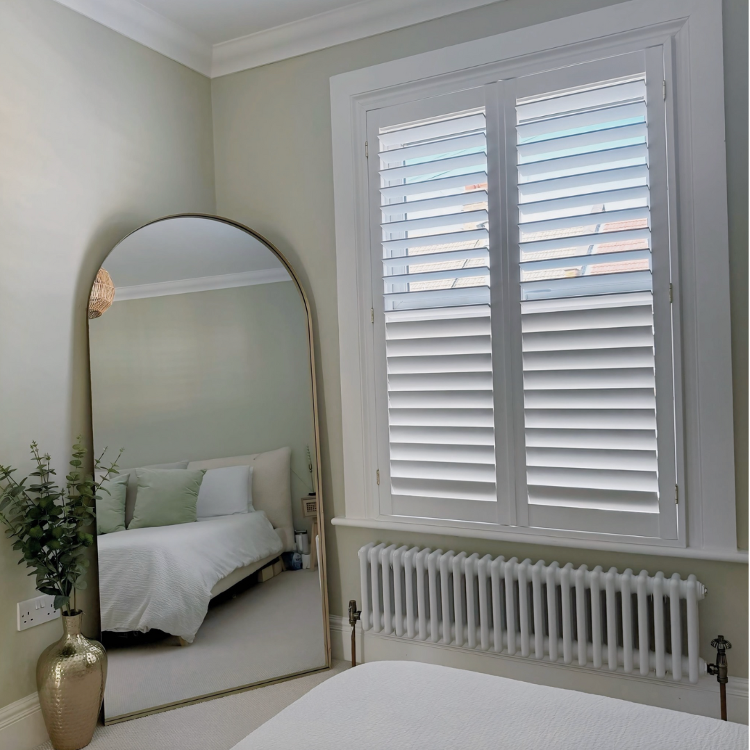 A bedroom with a large white-framed window with plantation shutters, a white bed with green pillows reflected in a tall, arched mirror, a radiator below the window, and a potted plant next to the mirror.