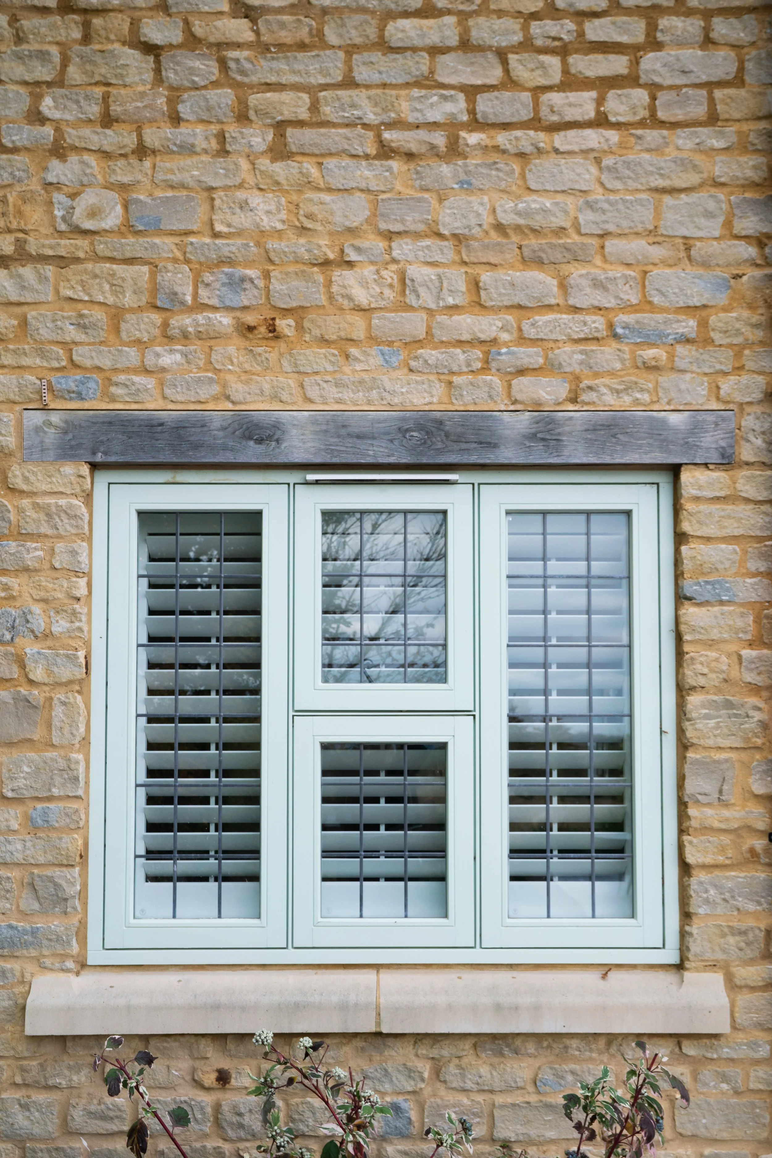 A window with white shutters and a wooden beam above, set into a beige stone brick wall with some plants below.