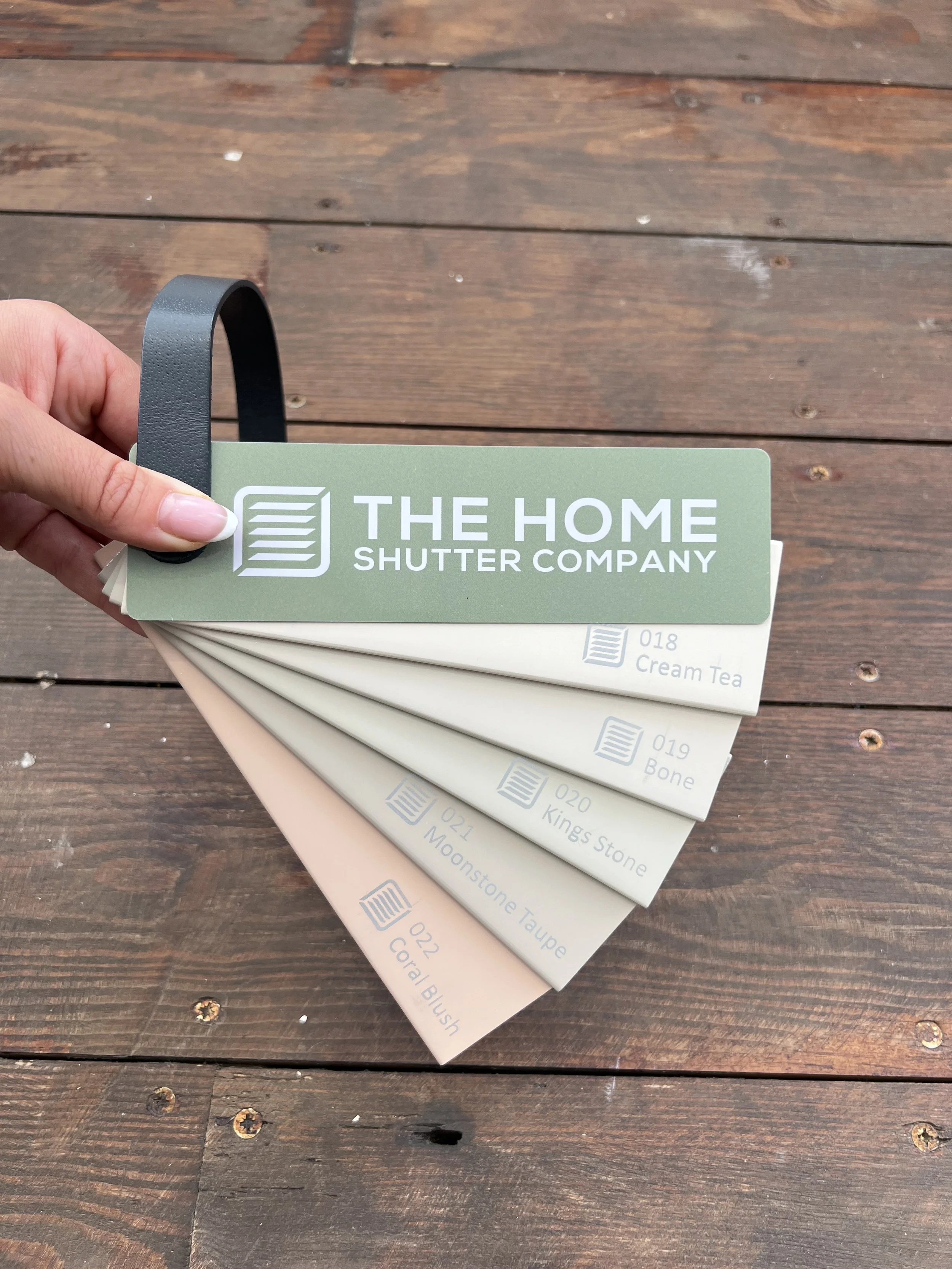 Hand holding a set of paint color sample cards from The Home Shutter Company, fanned out on a wooden surface.