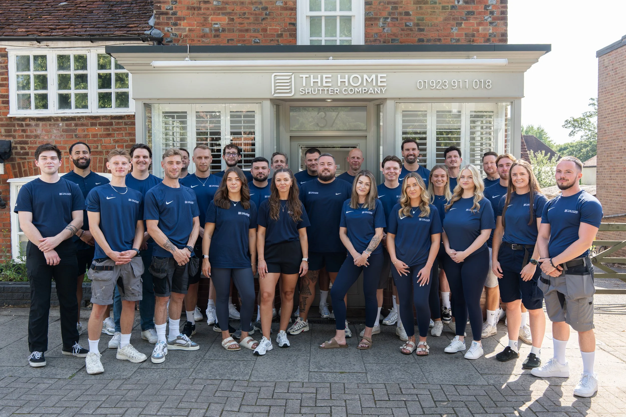 Group of people standing outside in front of a building with a sign that reads 'The Home Shutter Company.'