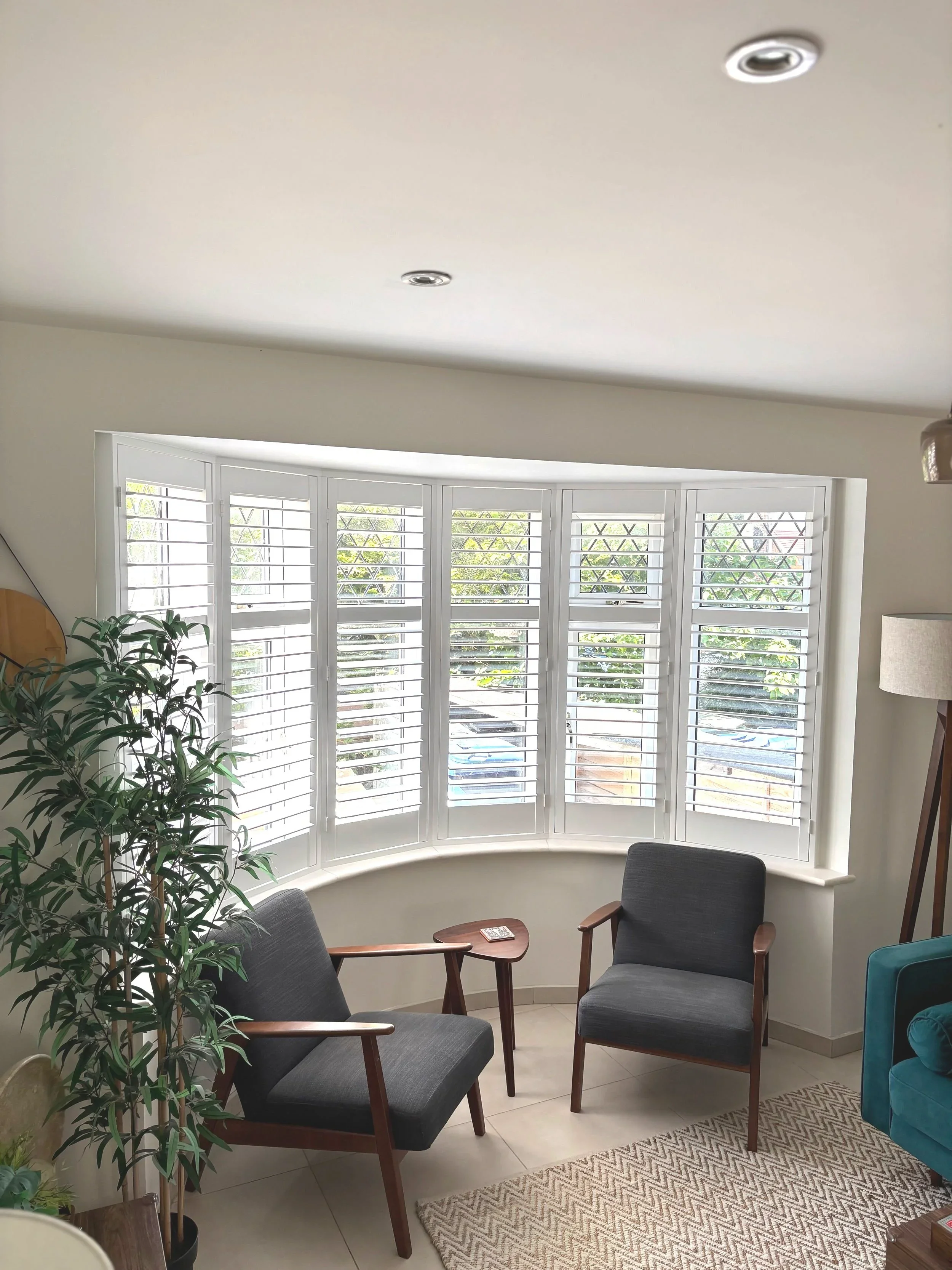 Living room with large bay window featuring white shutters, two gray armchairs with wooden arms, a side table, potted plant, a beige and white patterned rug.