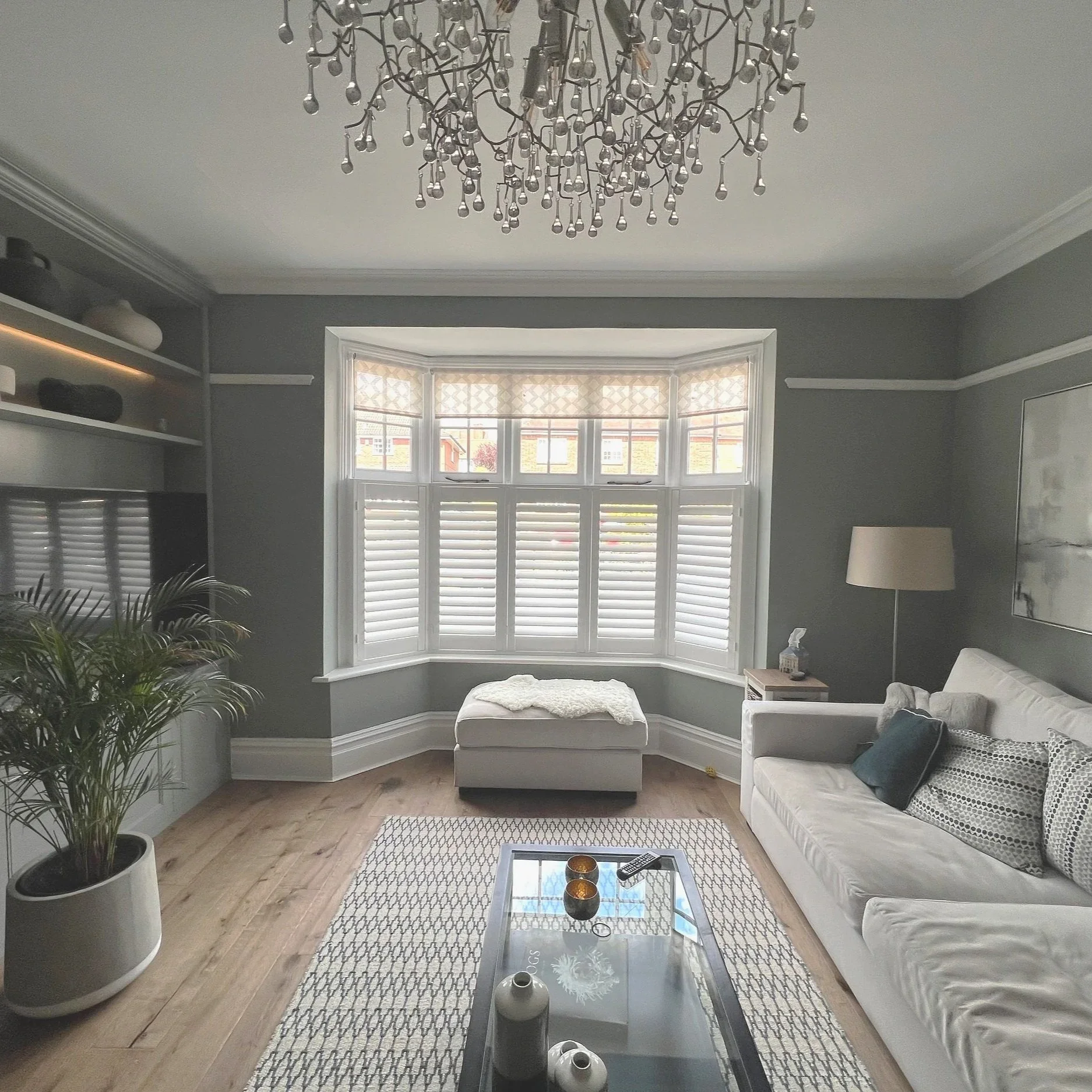 Living room with a large window, white sofa with pillows, a glass-top coffee table, a floor lamp, a rug, and a potted plant, with a chandelier hanging from the ceiling.