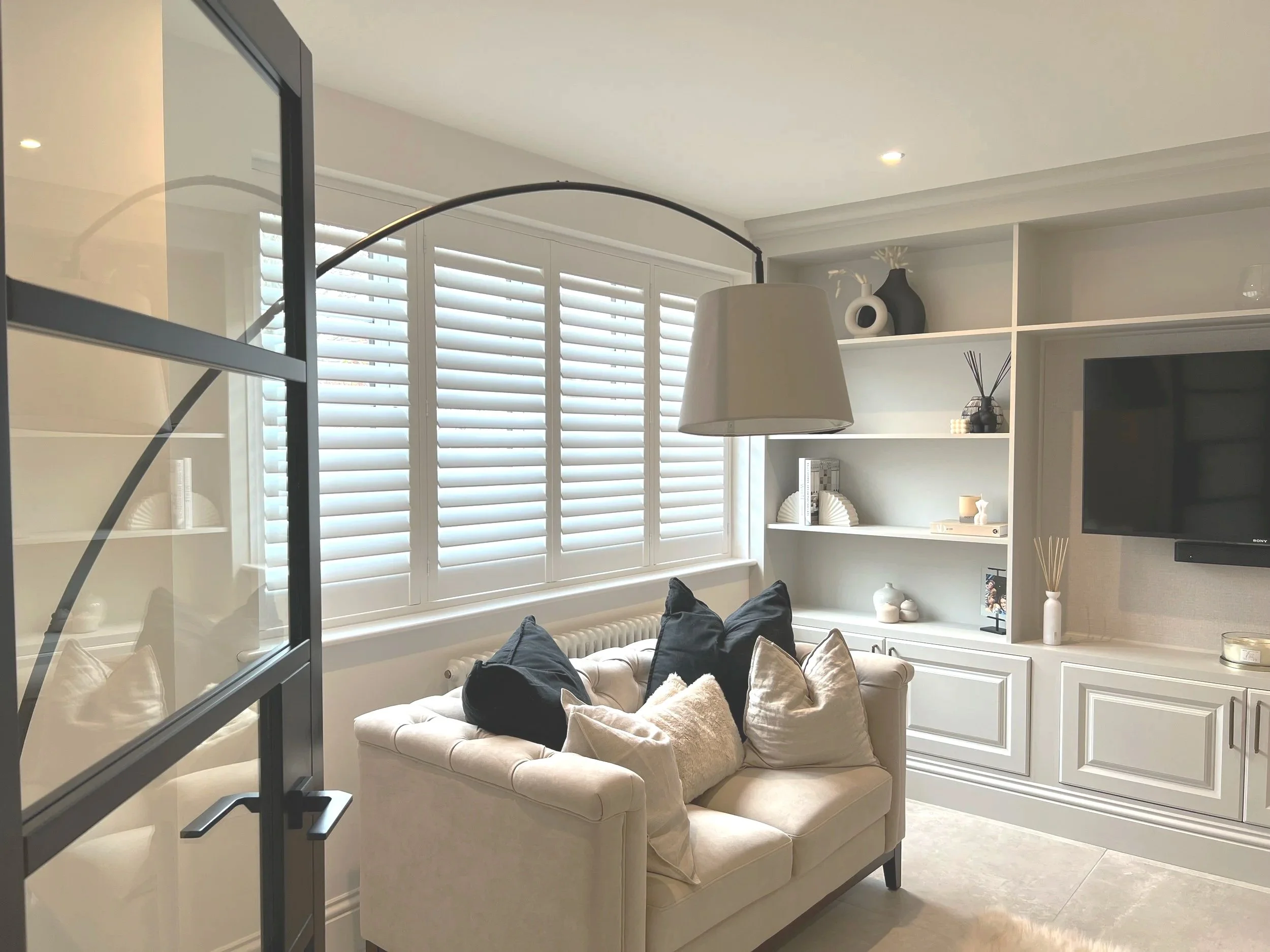 A living room with a large window covered with white plantation shutters, a beige tufted sofa with black and beige pillows, and a built-in white shelf with decorative vases and books. A large white floor lamp arches over the sofa, and a black TV is mounted on the wall.