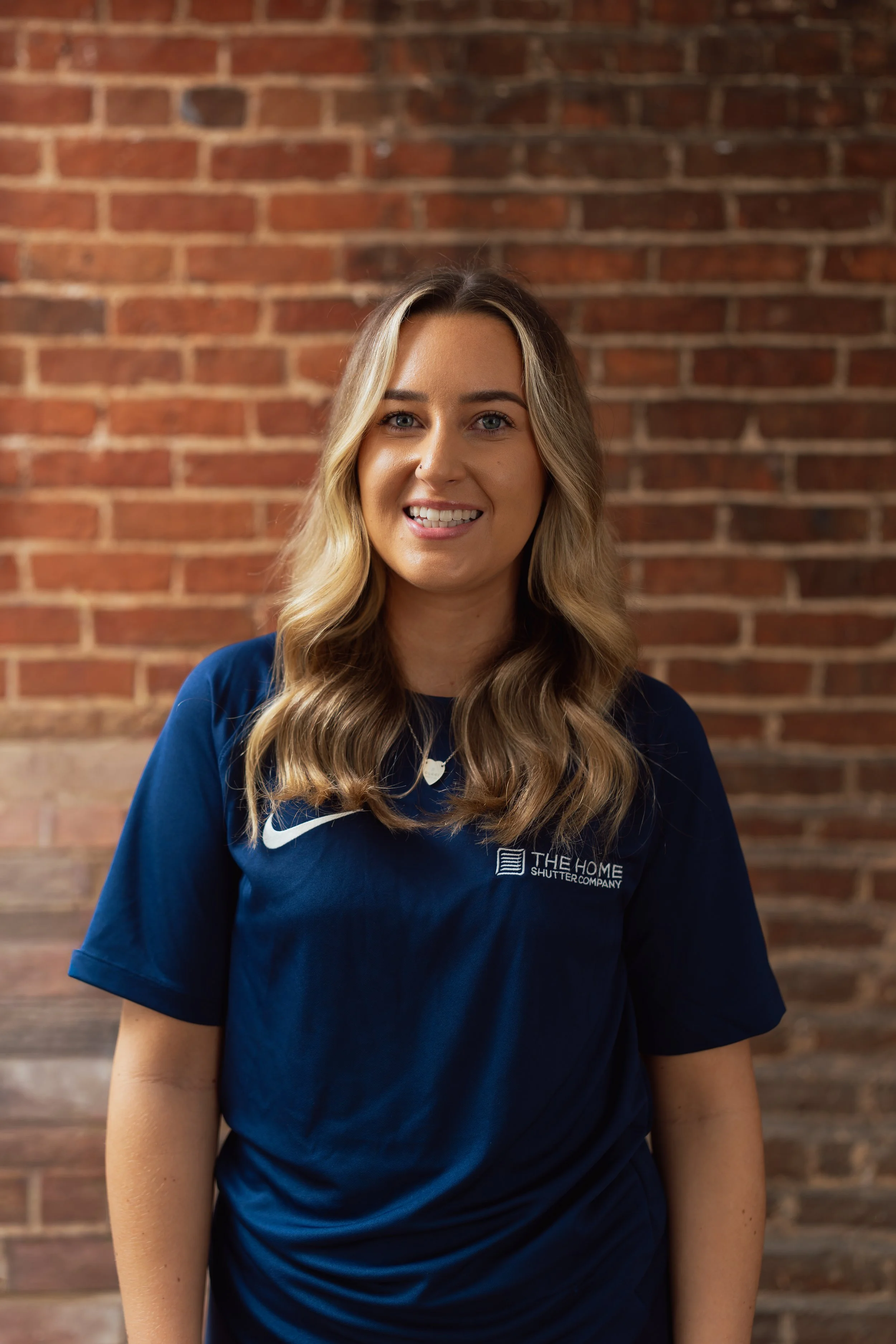 Portrait of a smiling young woman with long wavy blonde hair, wearing a navy blue t-shirt with logo, standing in front of a brick wall.