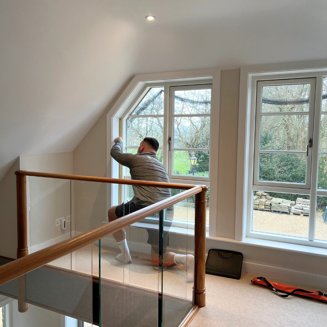 A man kneeling on a window seat cleaning or installing a window in a bright room with two large windows and a view of trees outside.