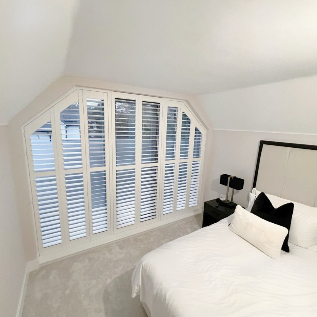 A bedroom with a large window with white plantation shutters, white walls, and a bed with white bedding and black and white pillows. There is a black nightstand with a black lamp and a remote control.