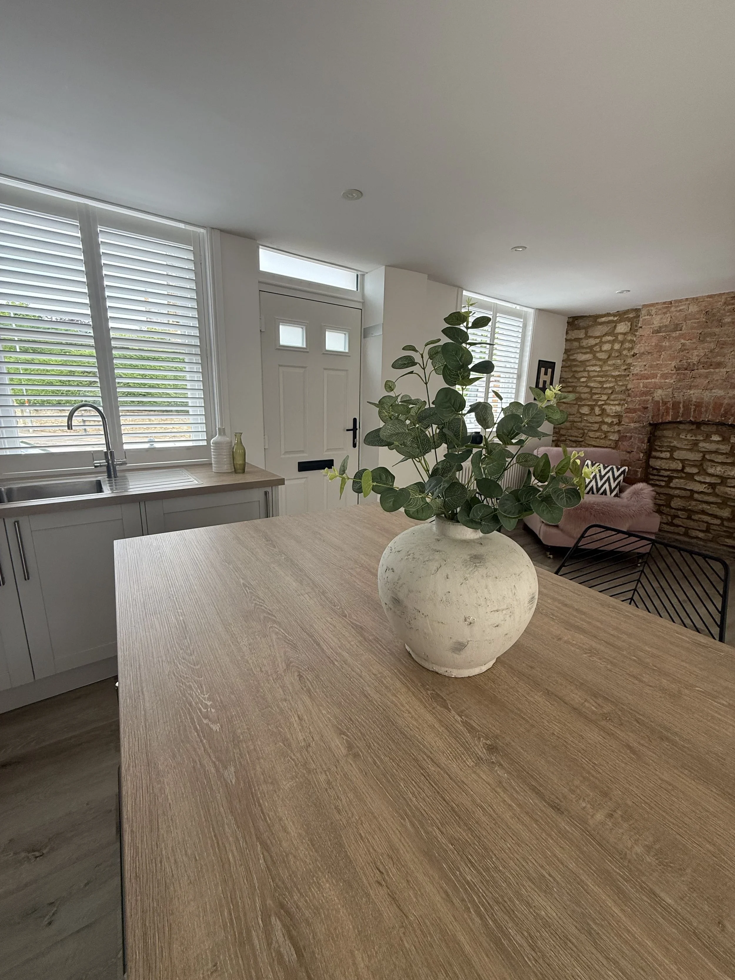 A modern kitchen with a wood dining table, a potted plant in a white textured vase, white cabinets, a sink, and large windows with white blinds, and a brick wall in the living area.