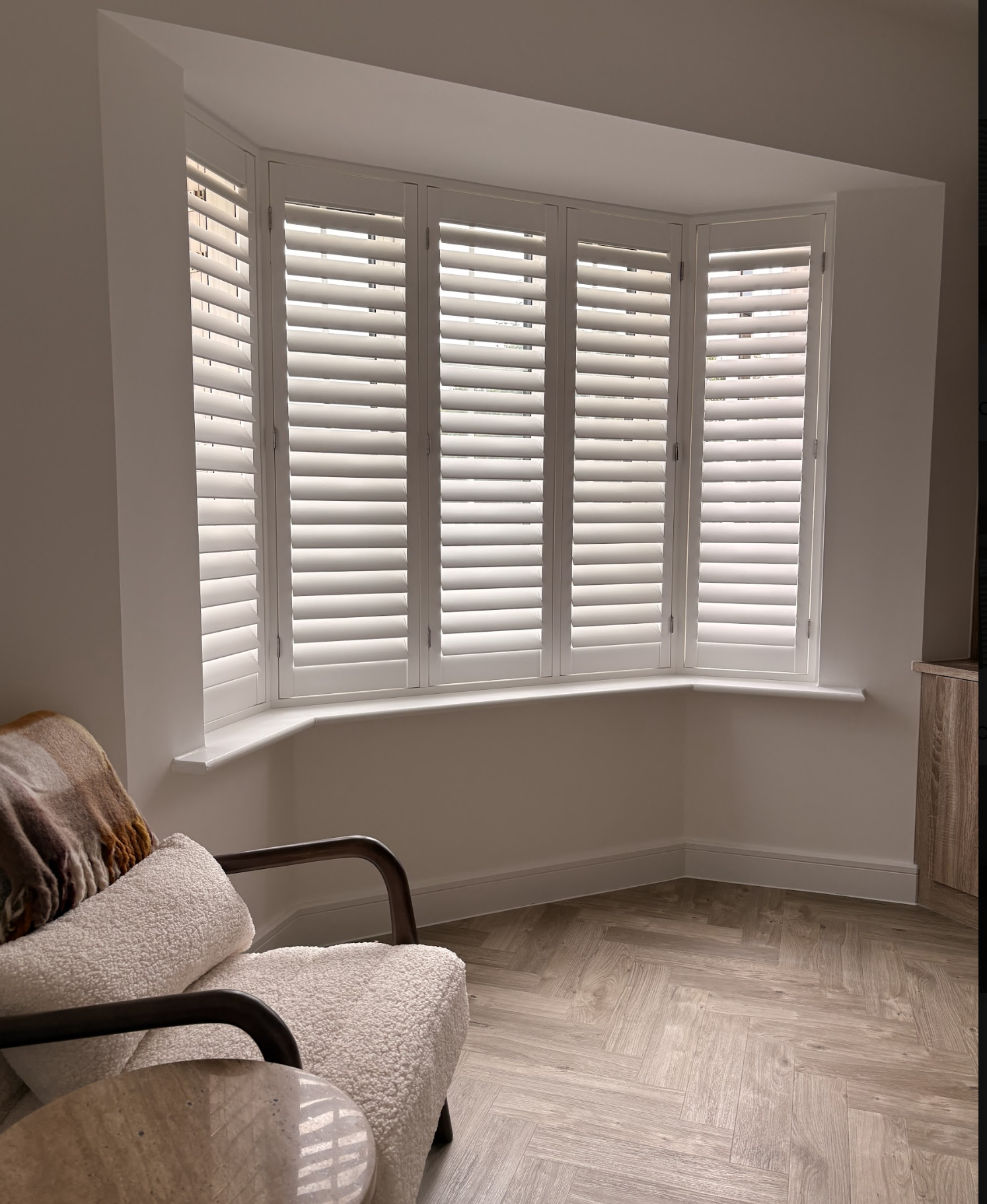 A room with a large bay window with white shutters, a beige armchair with a throw blanket, a wooden side table, and wood flooring.
