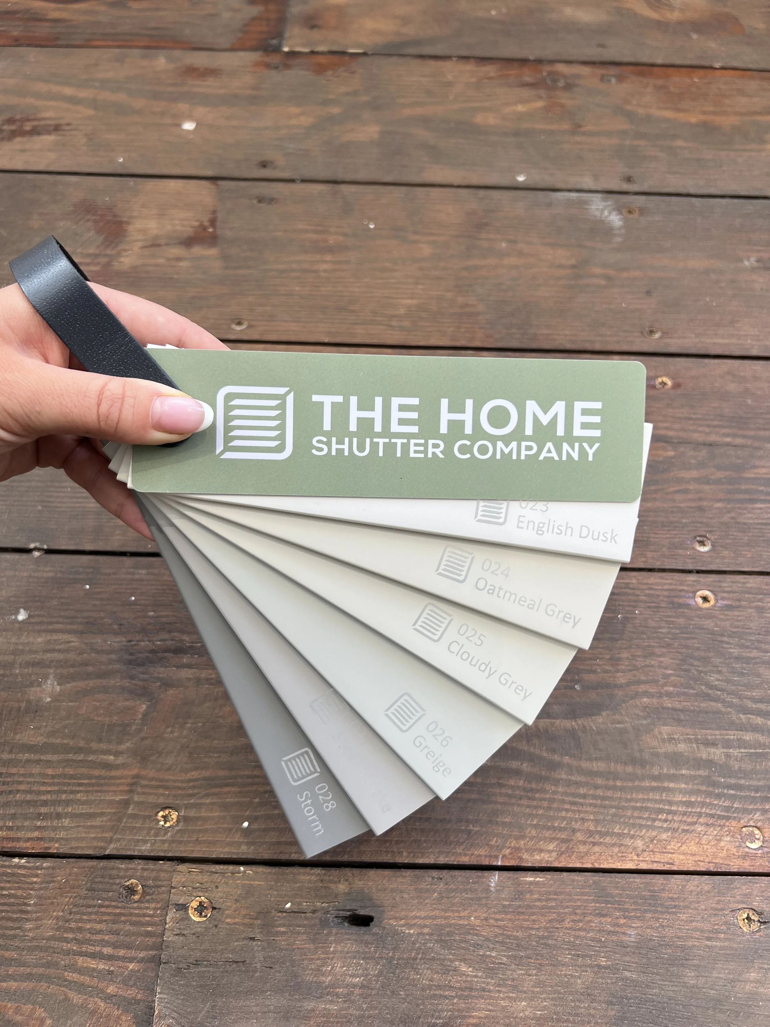 Hand holding color sample book from The Home Shutter Company, fanned out on a wooden surface.