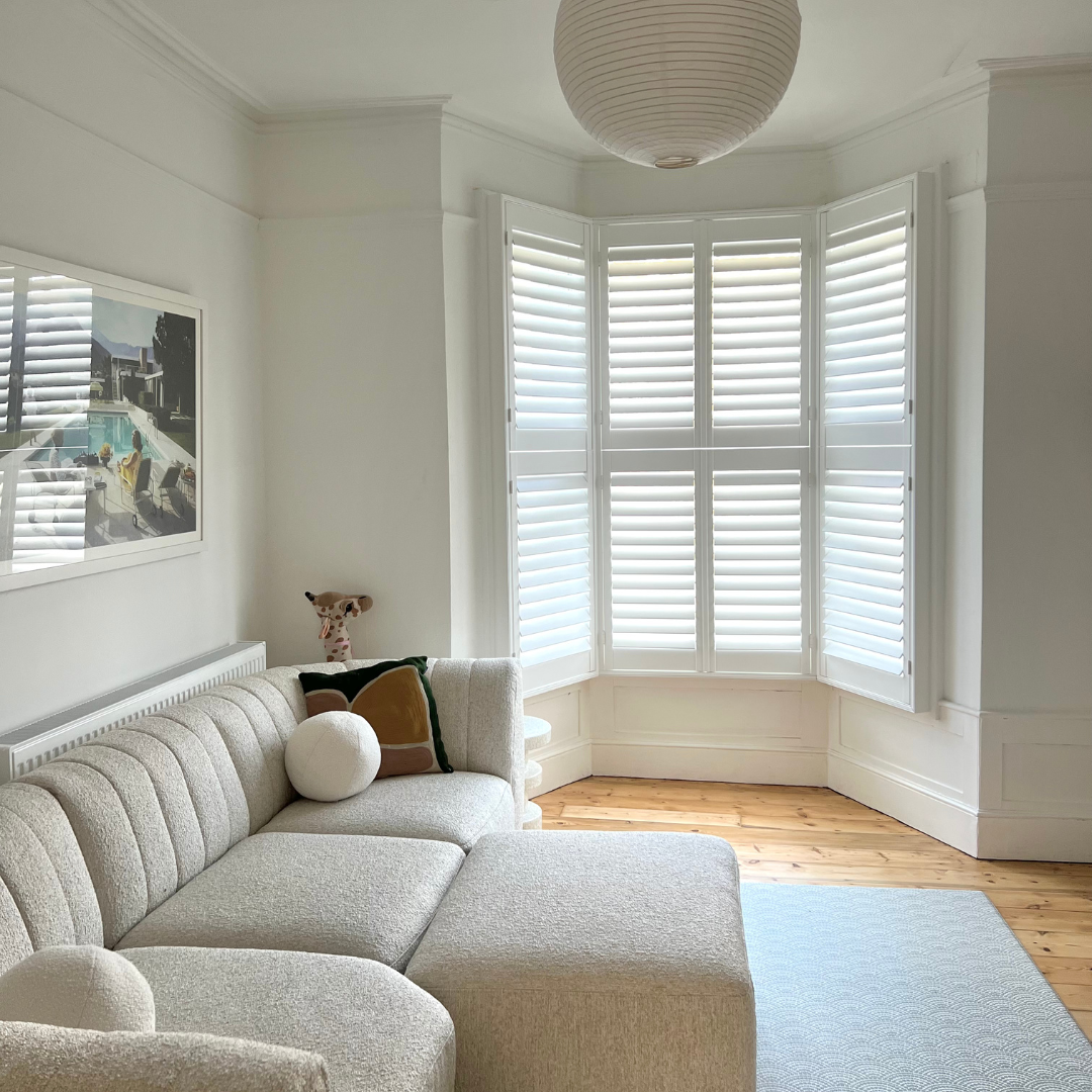 Living room with a large white sectional sofa, pillows, a lap blanket, a framed art piece on the wall, hardwood floors, and a bay window with white shutters.