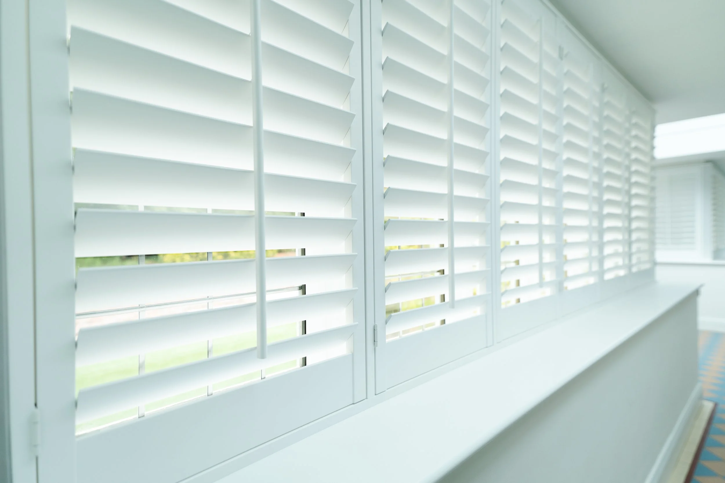 Close-up of white window shutters partially open, allowing light to come through, with a view of green trees outside.