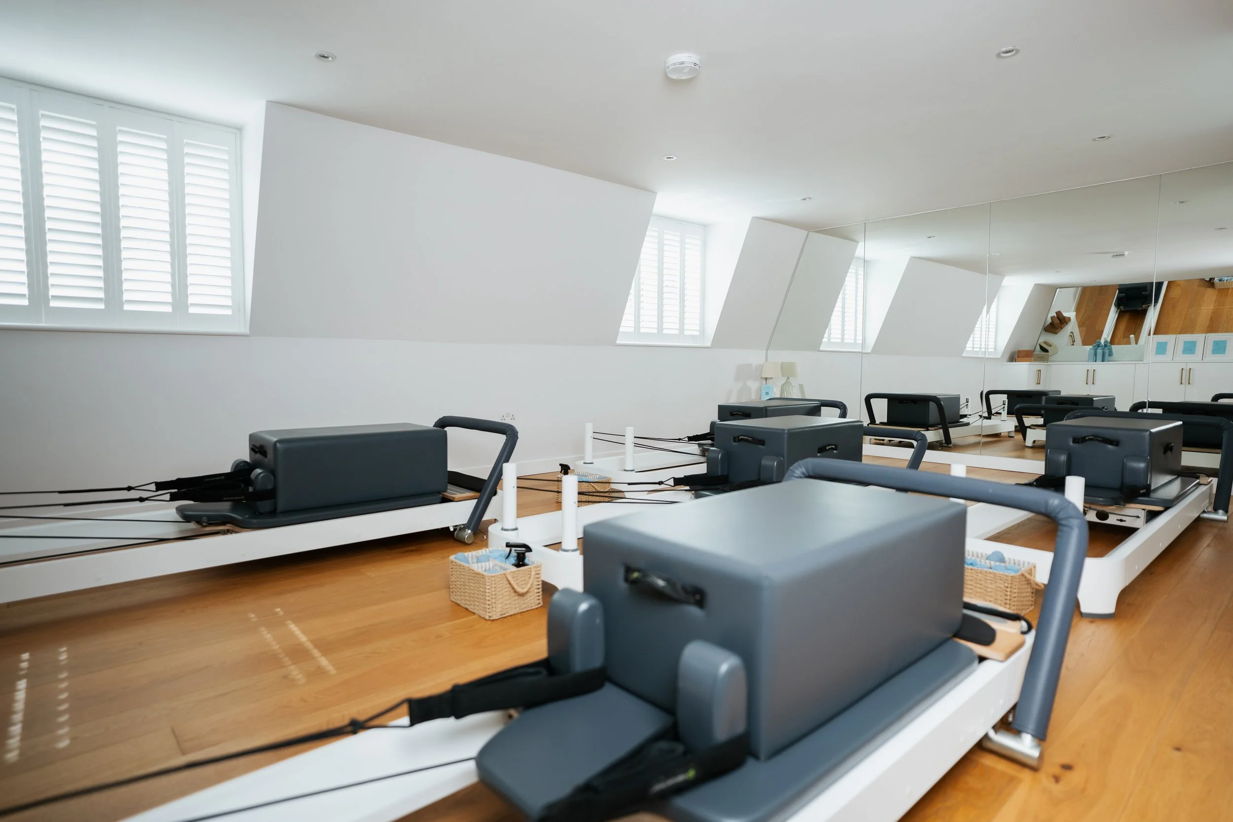 A fitness or physical therapy room with Pilates reformer machines arranged on a hardwood floor, large windows with white shutters, and a mirror on the wall reflecting the room's interior.