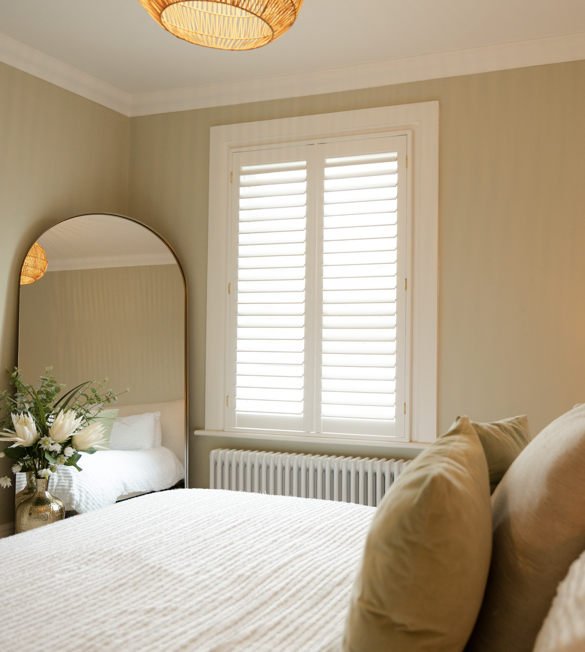 A bedroom with a bed, pillows, a nightstand with a vase of white flowers, a large window with white shutters, and a wall-mounted radiato