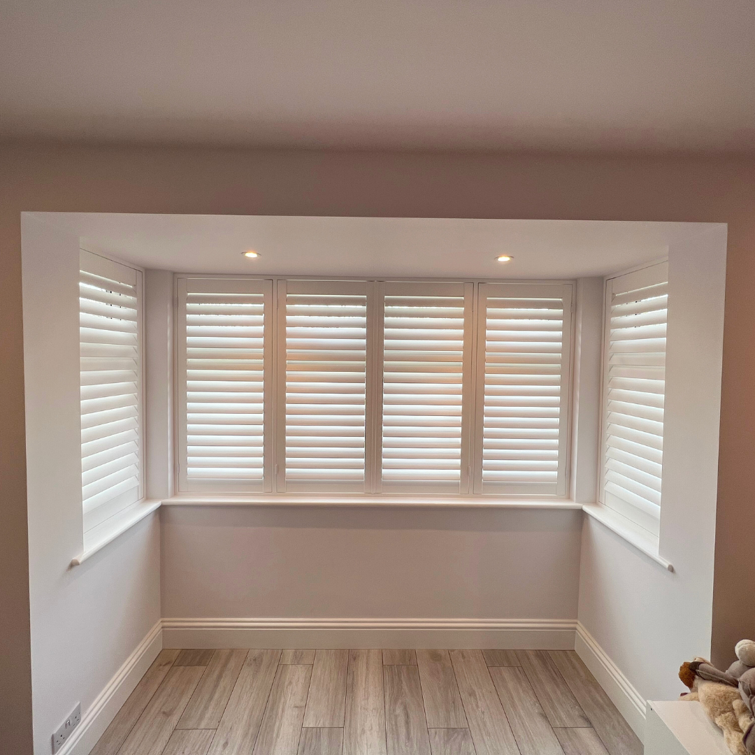 A room with three large windows covered by white shutters, beige walls, wooden flooring, recessed ceiling lights, and a white baseboard.
