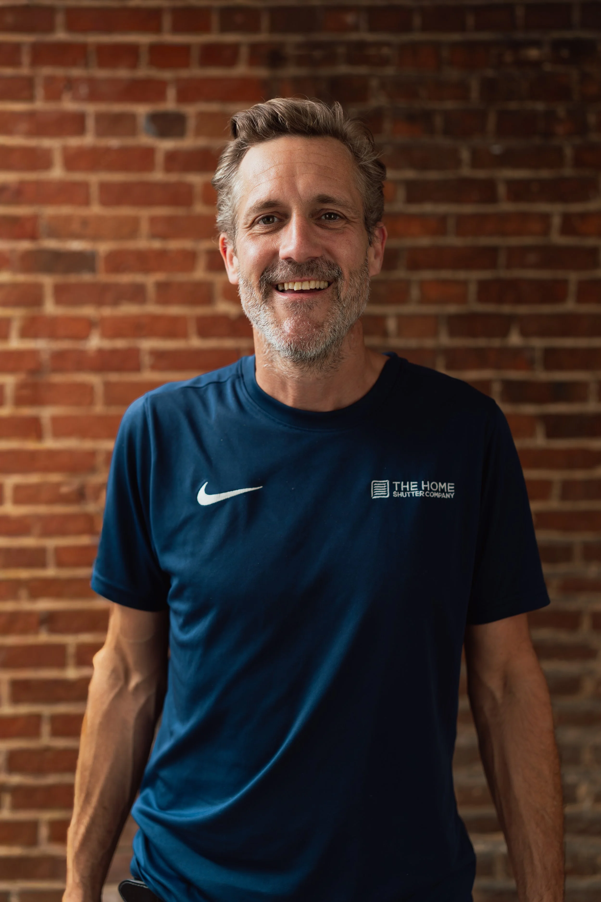 A middle-aged man with gray hair and beard smiling, wearing a navy blue Nike t-shirt with a logo, standing in front of a brick wall.