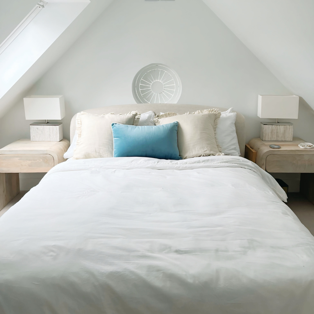 A neatly made bed with white sheets, a blue pillow, and beige pillows in a bright, minimalist bedroom with two bedside tables and lamps.