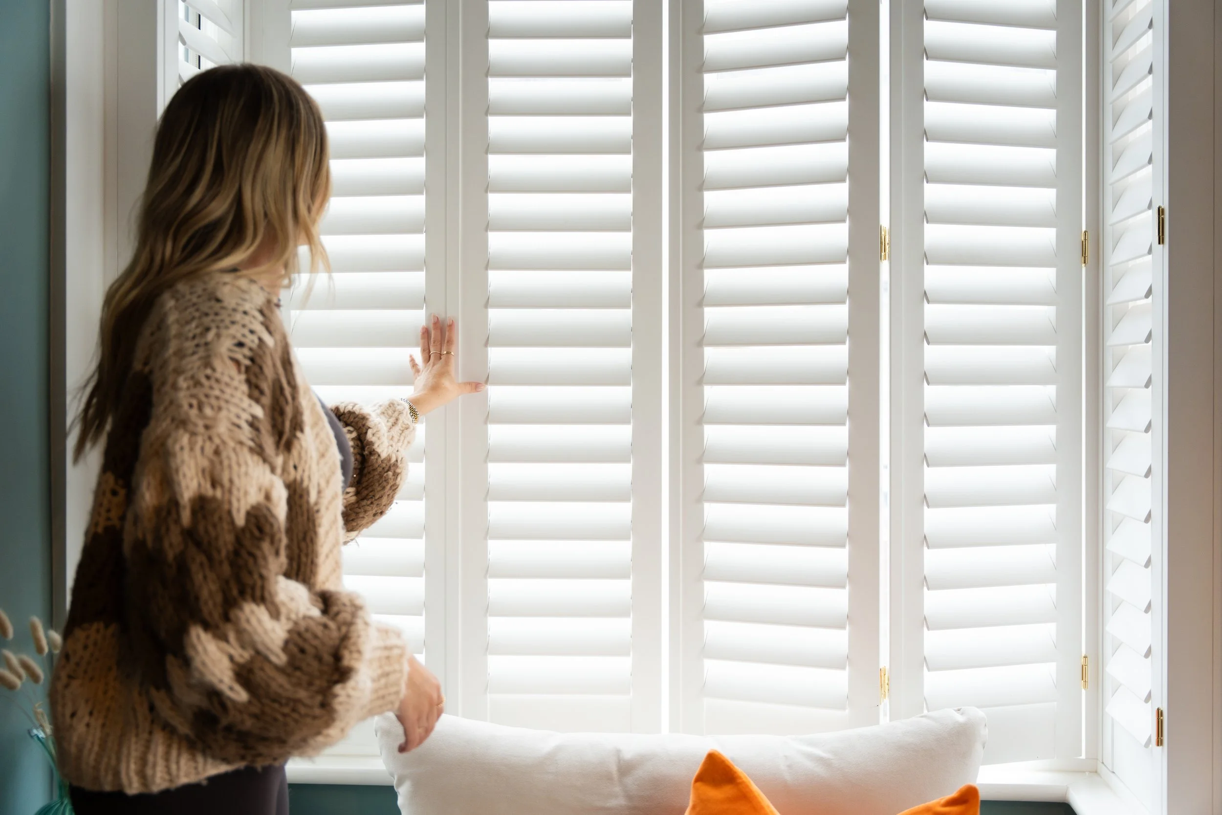 A woman with long brown hair wearing a brown and beige patterned sweater looking out of white plantation shutters.