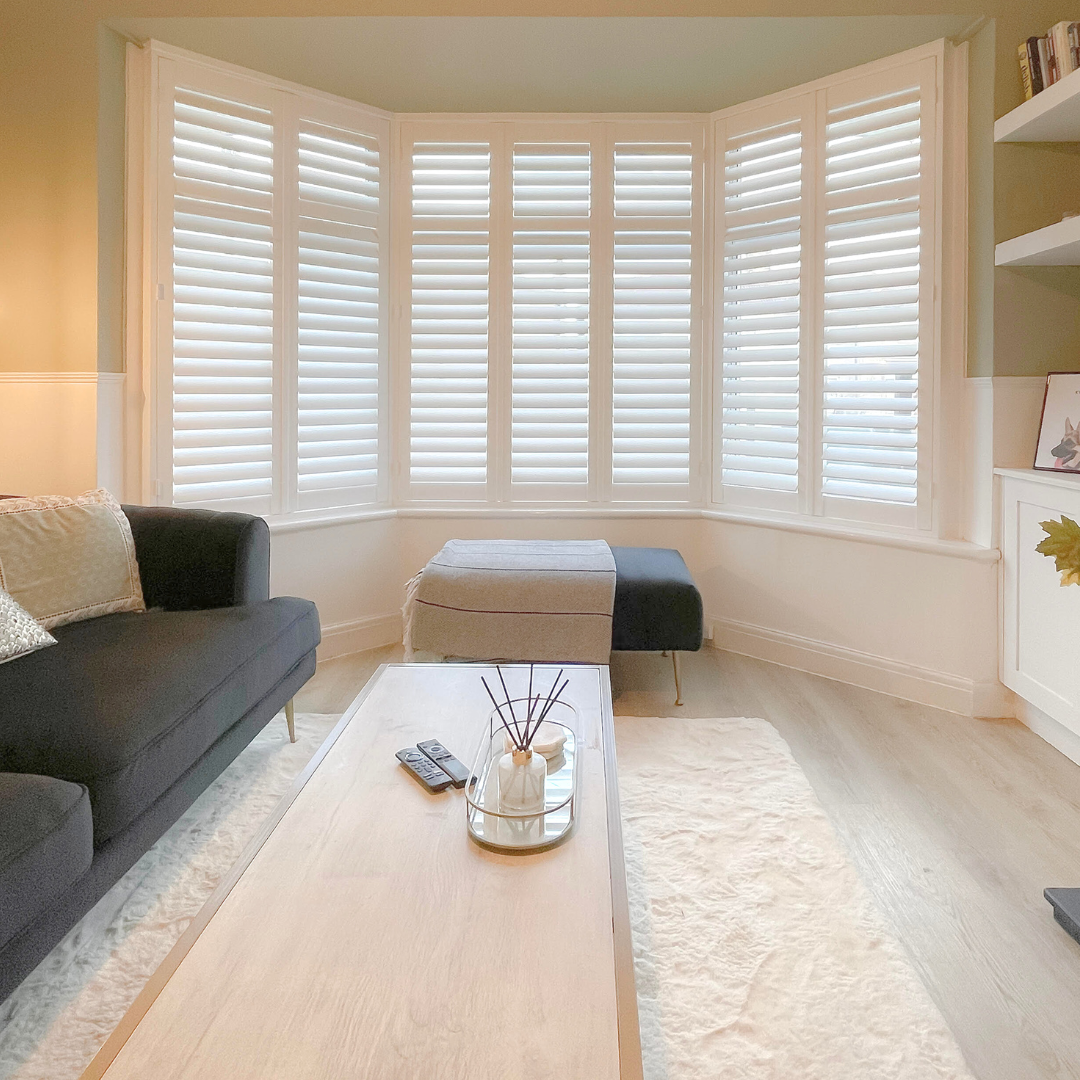 Living room with a large bay window with white plantation shutters, black sofa with beige and silver pillows, light wood coffee table with a decorative tray and remote control, cream rug, and a beige storage bench with a blanket on top.