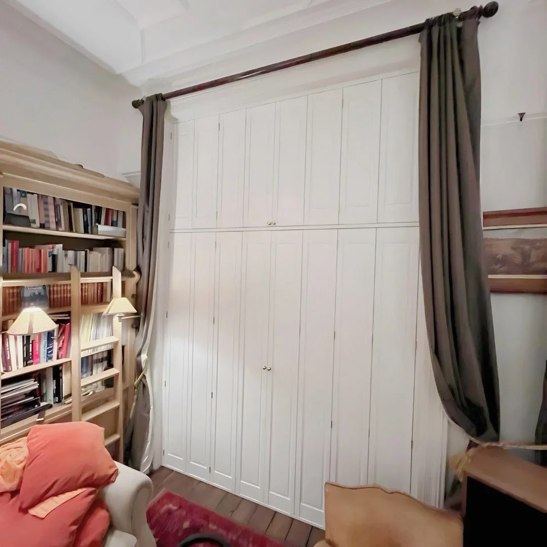 A room with white built-in closet with paneled doors, dark curtains on a brass curtain rod, a wooden bookshelf with lamps, a beige armchair with orange and yellow pillows, and part of a wooden desk, all on a hardwood floor with a red rug.