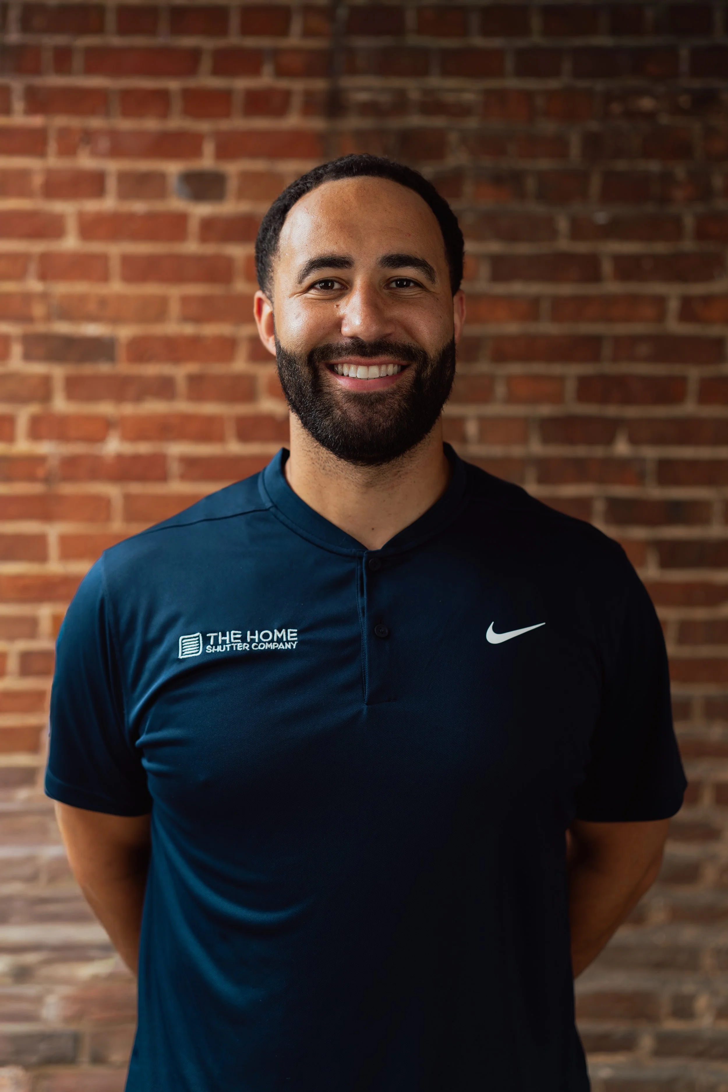 A man with a beard and short hair standing in front of a brick wall, wearing a navy blue Nike sports shirt with a logo on the left side and a company logo on the right side.