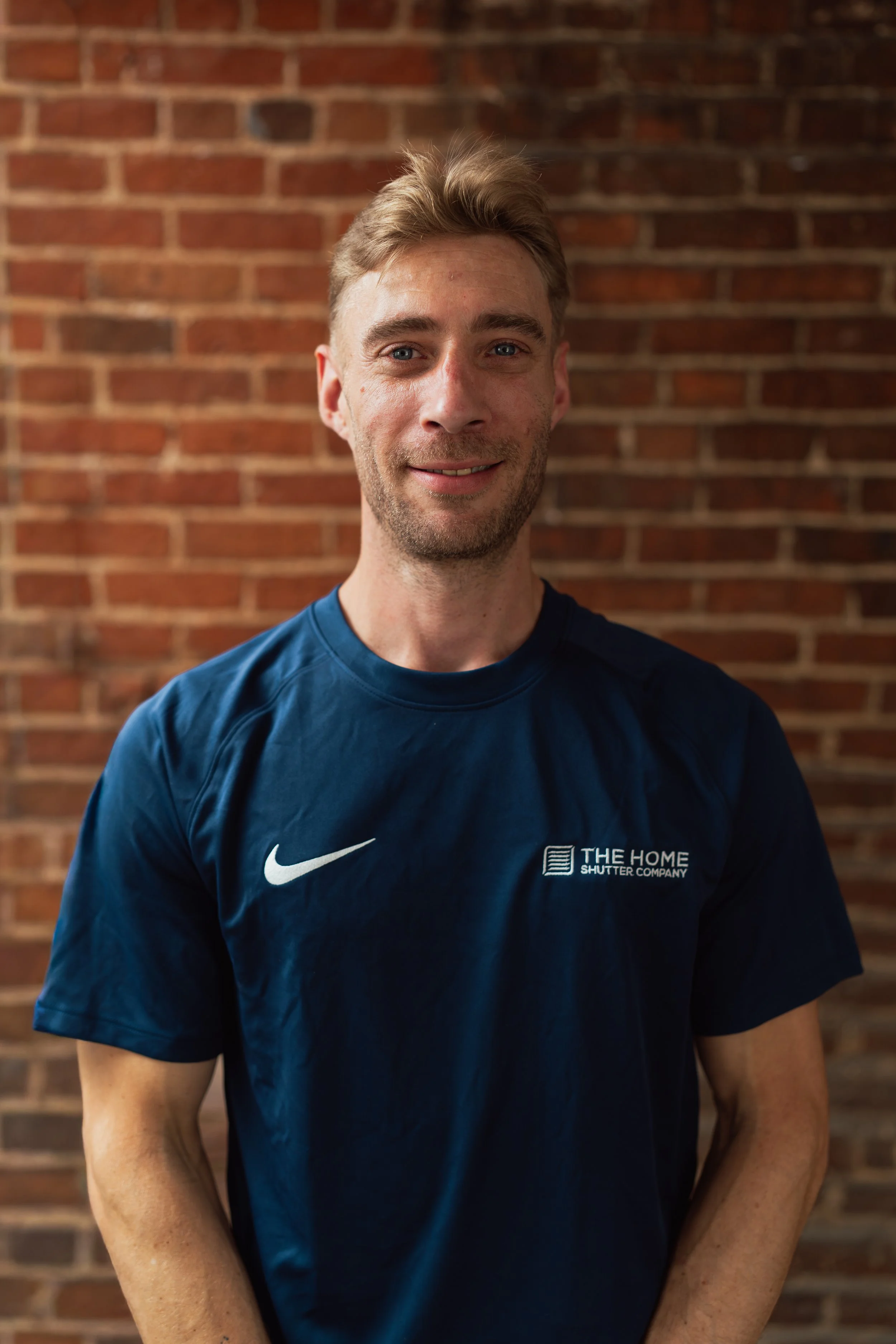 A man with short blond hair and blue eyes smiling while wearing a blue Nike athletic shirt against a brick wall background.