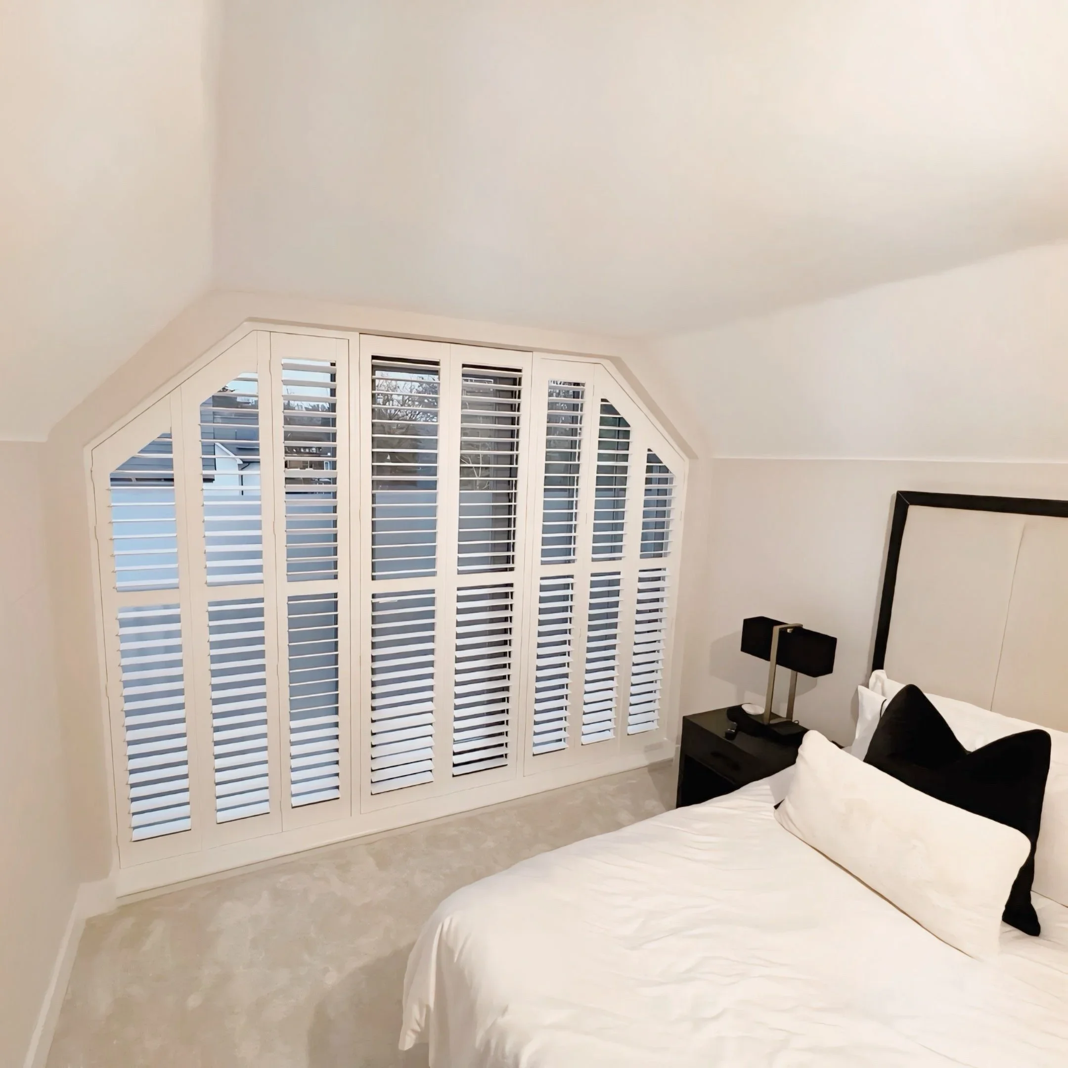 A bedroom with white walls, a white bed with black and white pillows, and a black bedside table with a black lamp. There is a large window with white plantation shutters and a view outside.