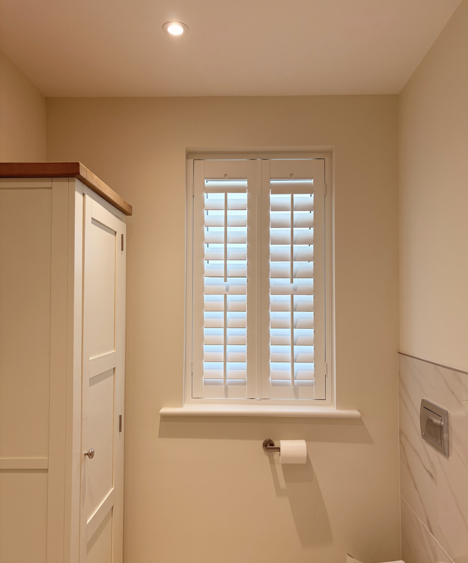 A bathroom with a window covered by white blinds, a white cabinet with a wooden top, a toilet paper roll on a holder, a wall-mounted silver switch, and beige walls with a recessed ceiling light.
