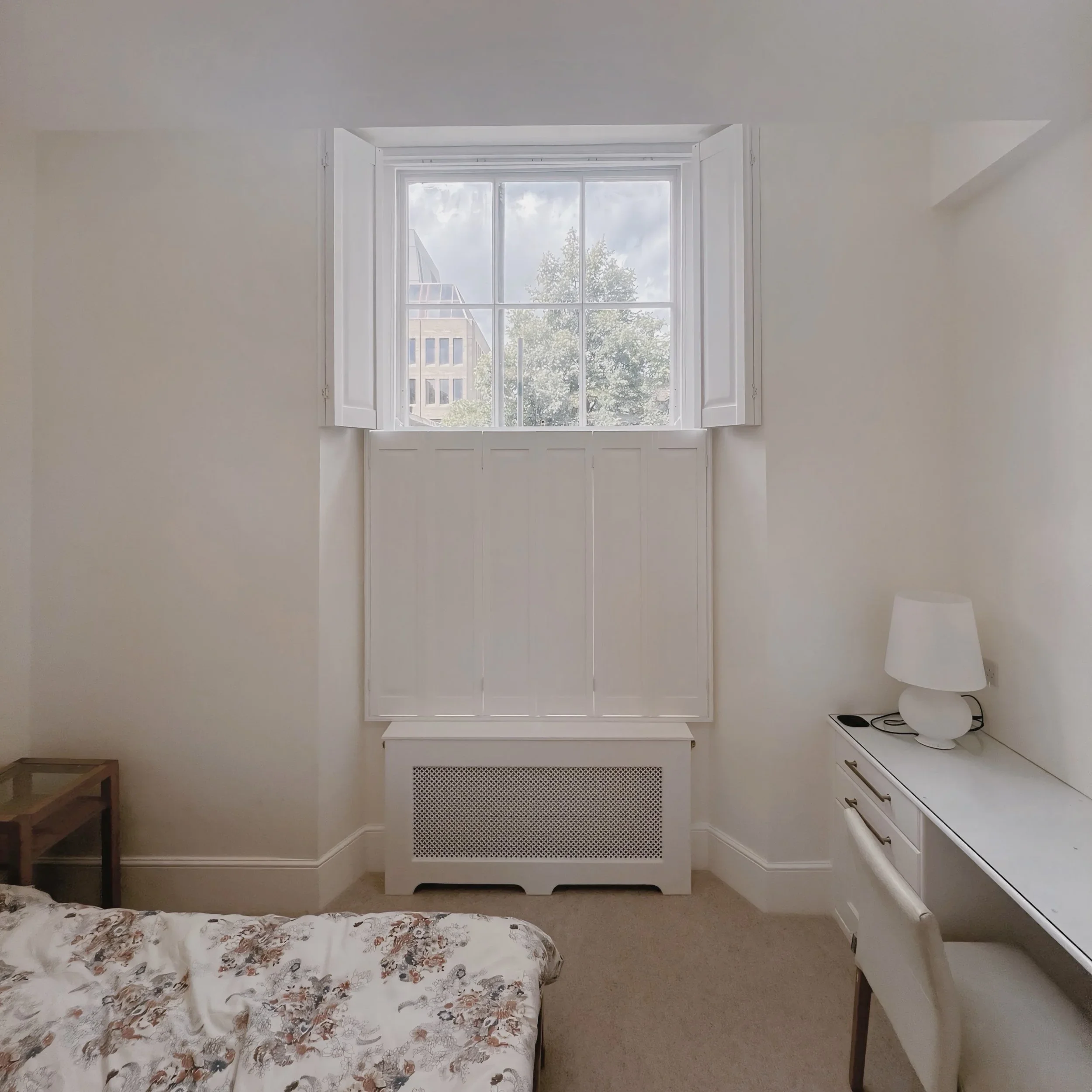 Room with a large window showing a tree and buildings outside, a bed with floral bedspread, a small bedside table, a white dresser, and a white lamp with a curved stand with tier on tier shutters top open
