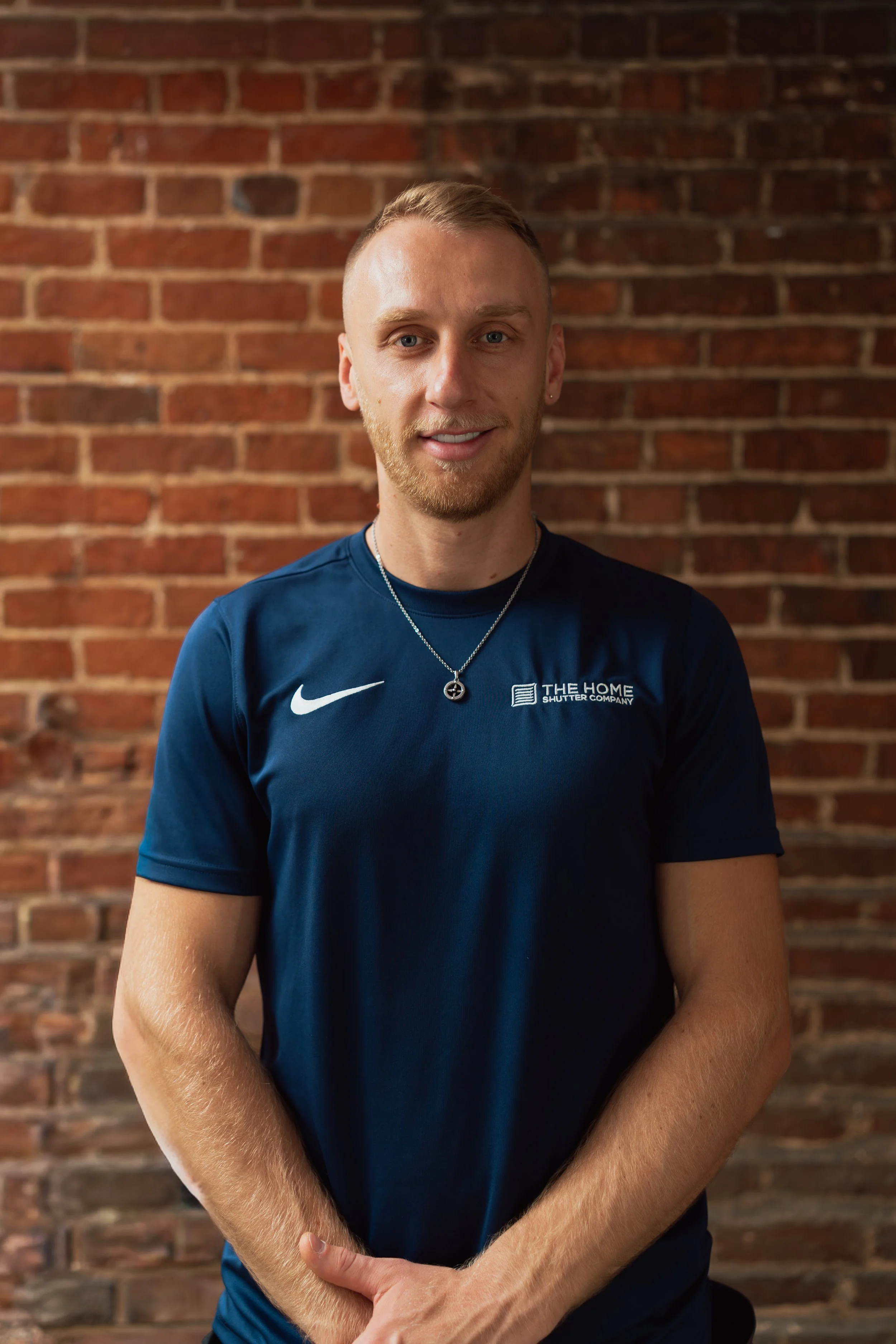 Portrait of a young man with short blonde hair and a beard, smiling slightly, wearing a navy blue Nike shirt with a logo and text, standing against a red brick wall.
