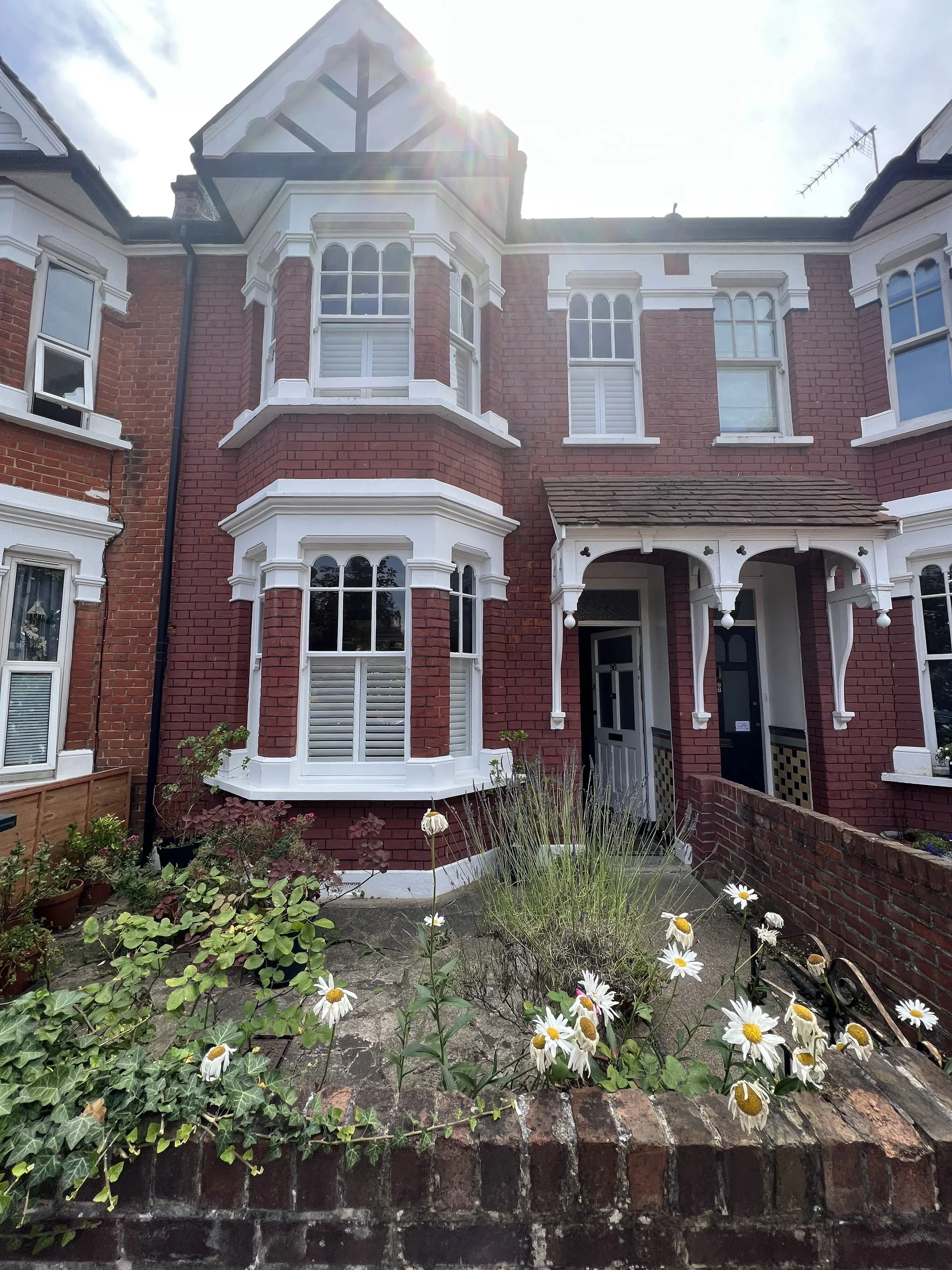 Red-brick Victorian house with white trim and bay windows, small garden with daisies and green plants, and a brick wall in the foreground.