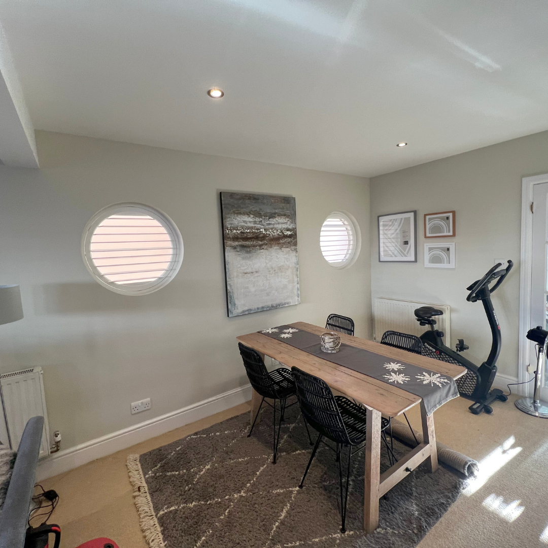 Dining area with wooden table, six black chairs, wall art, two small round windows, exercise bike, and rug on beige carpet.