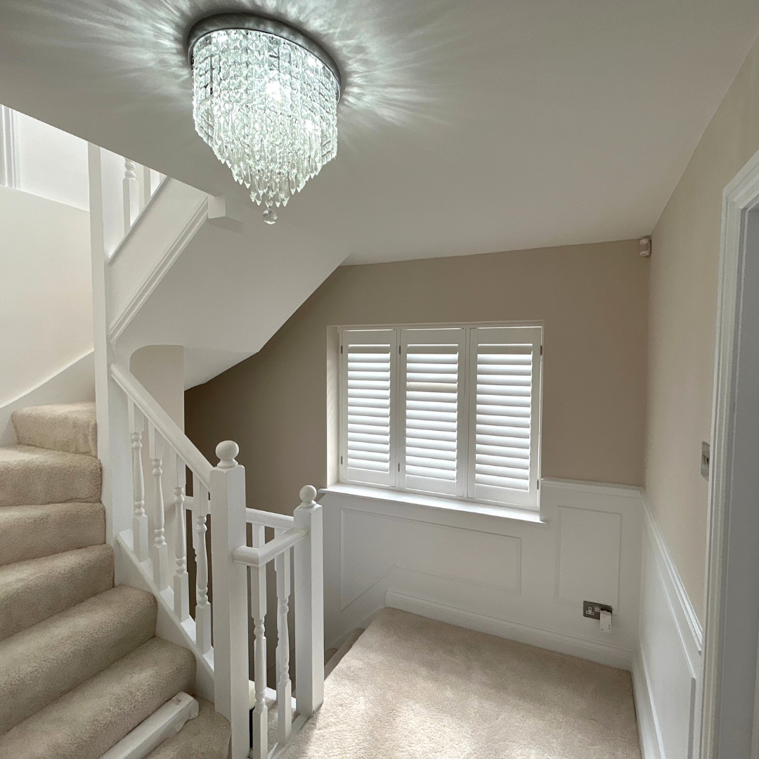 Interior view of a staircase landing with beige carpet, a white wooden railing, a window with white shutters, and a chandelier hanging from the ceiling.