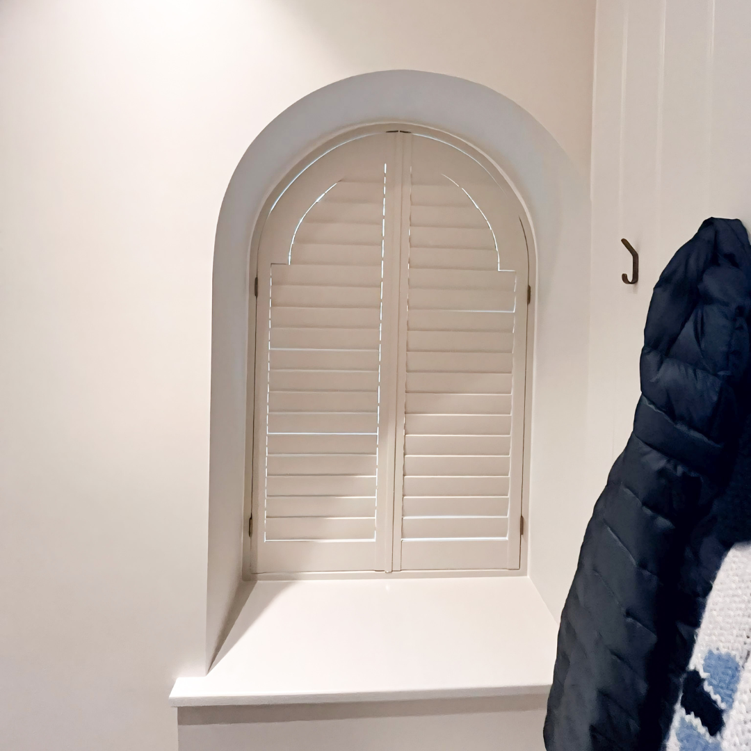White arched window with closed shutters and a window seat beneath it, in a room with white walls and a black coat hanging on a hook.