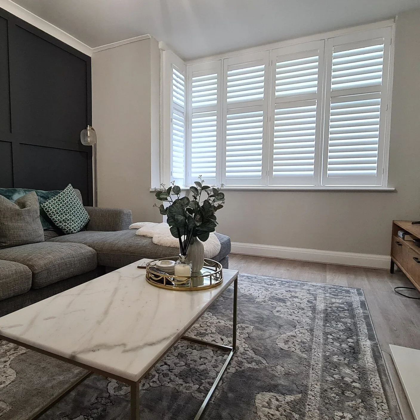 Living room with gray sectional sofa, marble coffee table with decorative tray and plant, large window with white shutters, neutral wall colors, hardwood floor, and a wooden TV stand.
