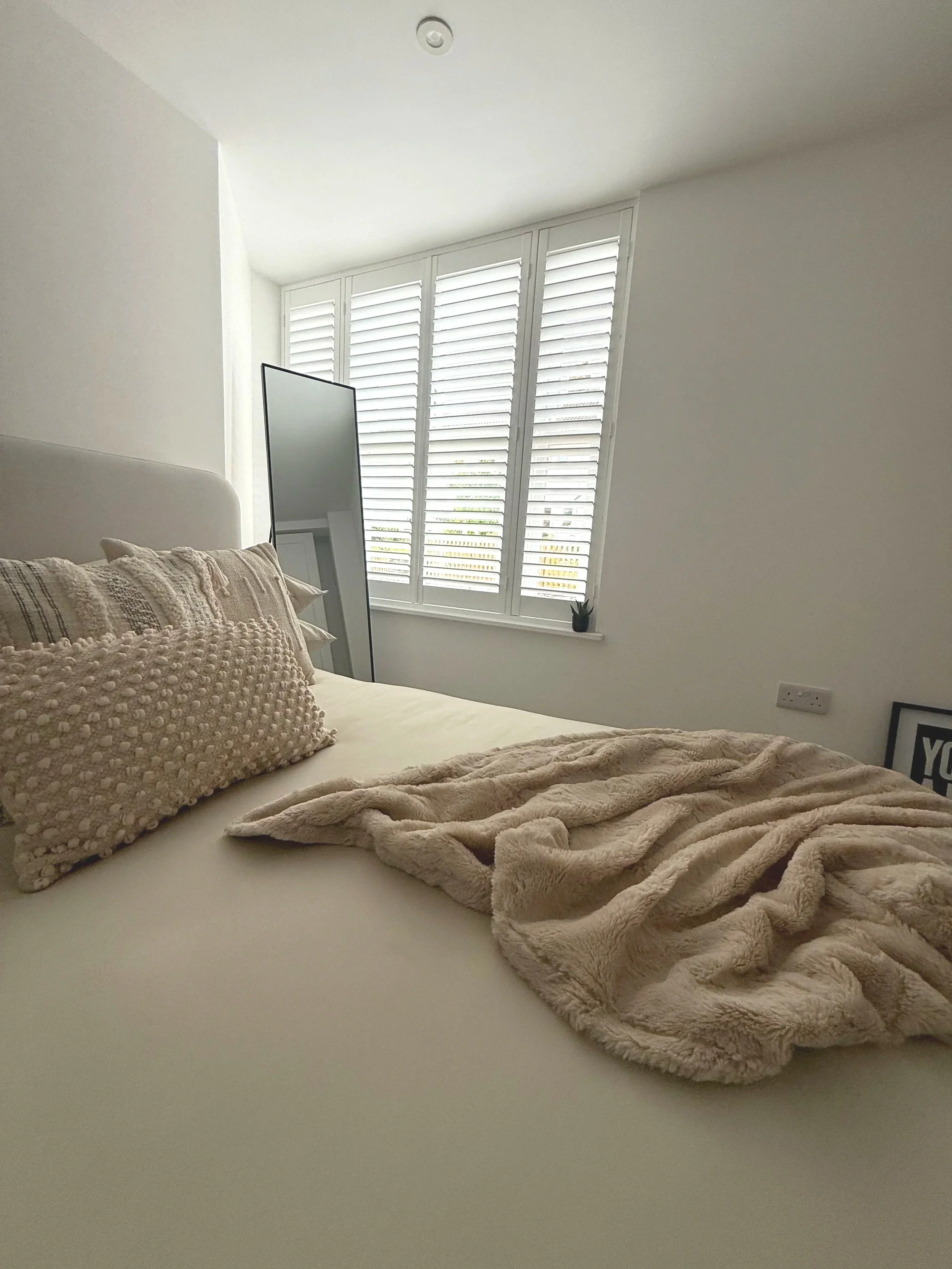 A cozy bedroom with a bed, decorative pillows, a faux fur throw blanket, a mirror, large window with wooden blinds, and a small plant on the window sill.