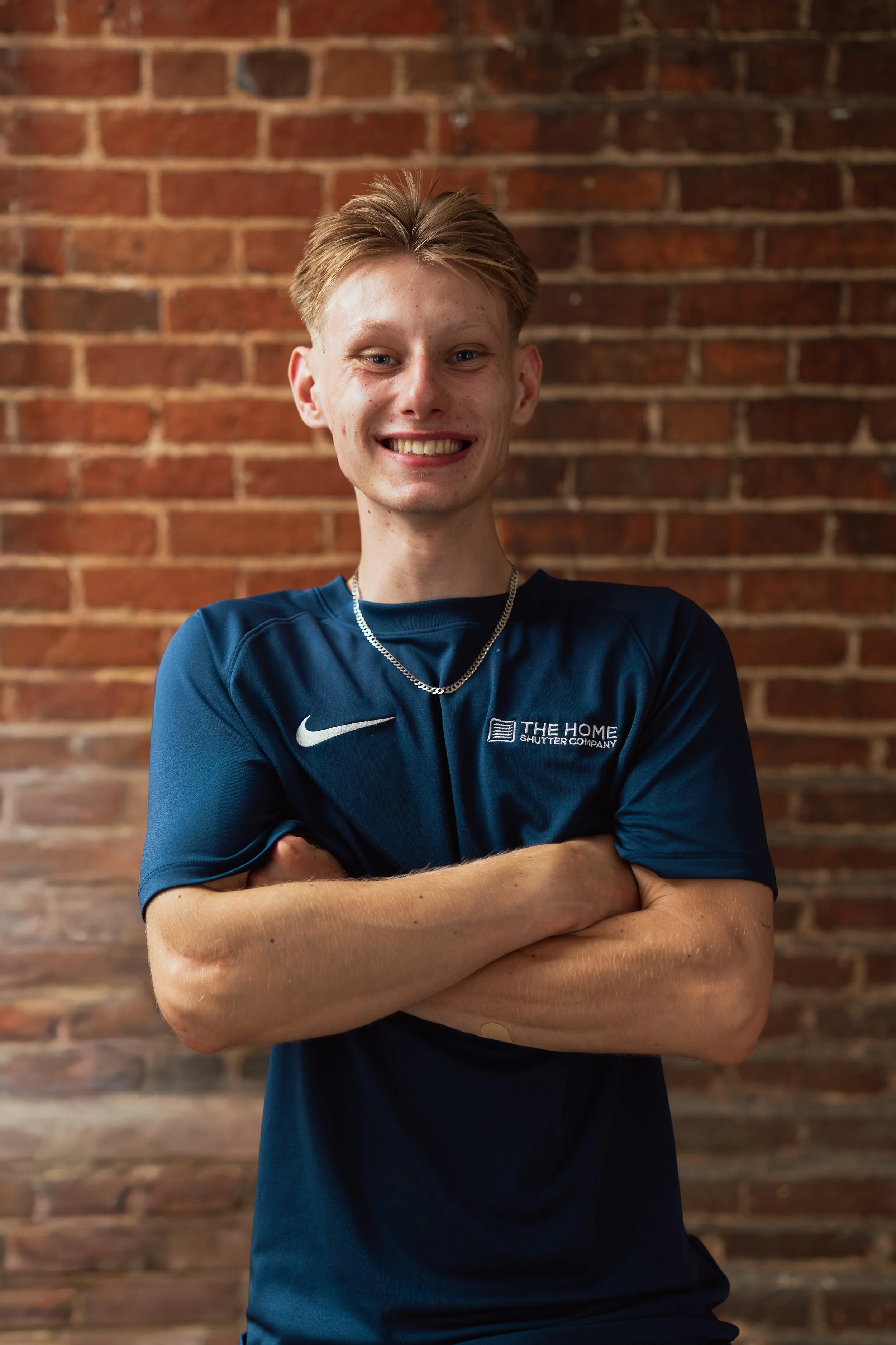 A young man with short, reddish hair and blue eyes standing in front of a brick wall, smiling with arms crossed, wearing a blue sports shirt with a logo on the chest and a silver chain necklace.