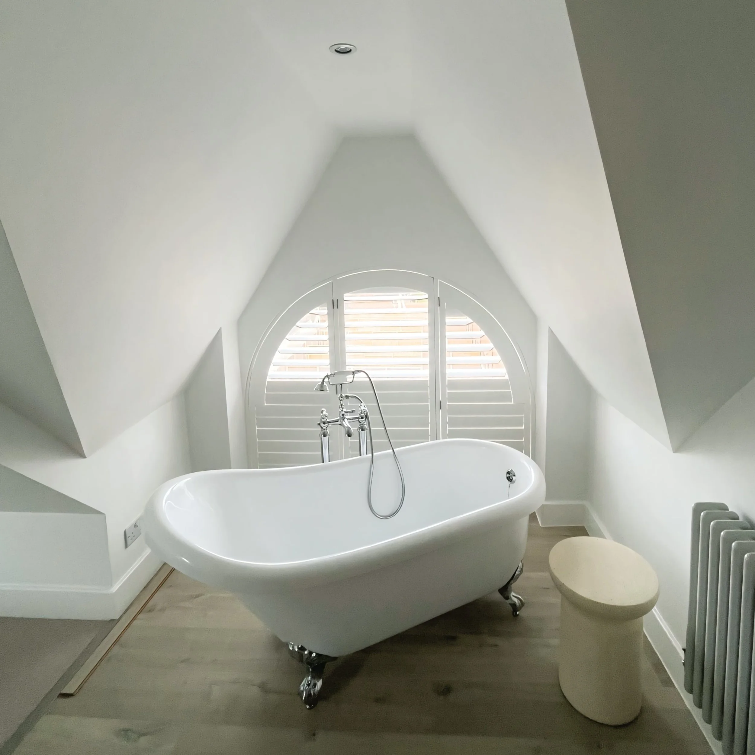White bathtub with a silver faucet and hand-held showerhead in front of a large arched window with white shutters, in a bathroom with white walls and gray flooring.