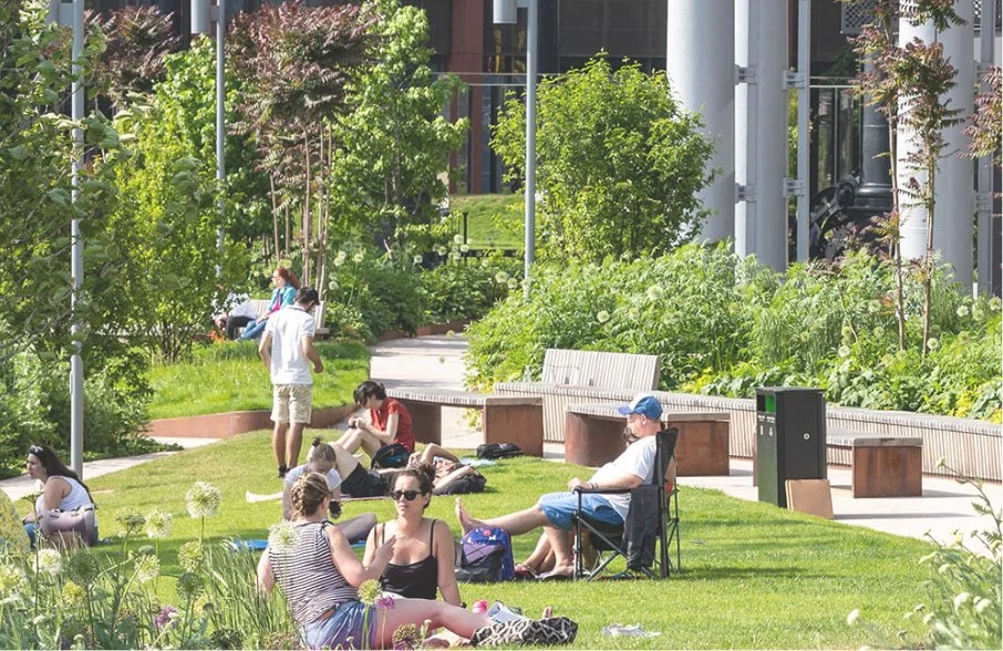 People relaxing and socializing in a lush, green park with trees, bushes, and garden beds, benches, and modern buildings in the background.