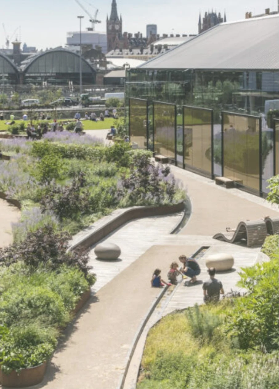 A park with greenery and a winding path, where a woman and three children are sitting on the ground. In the background, people are sitting on benches and walking, with city buildings and a glass structure nearby.