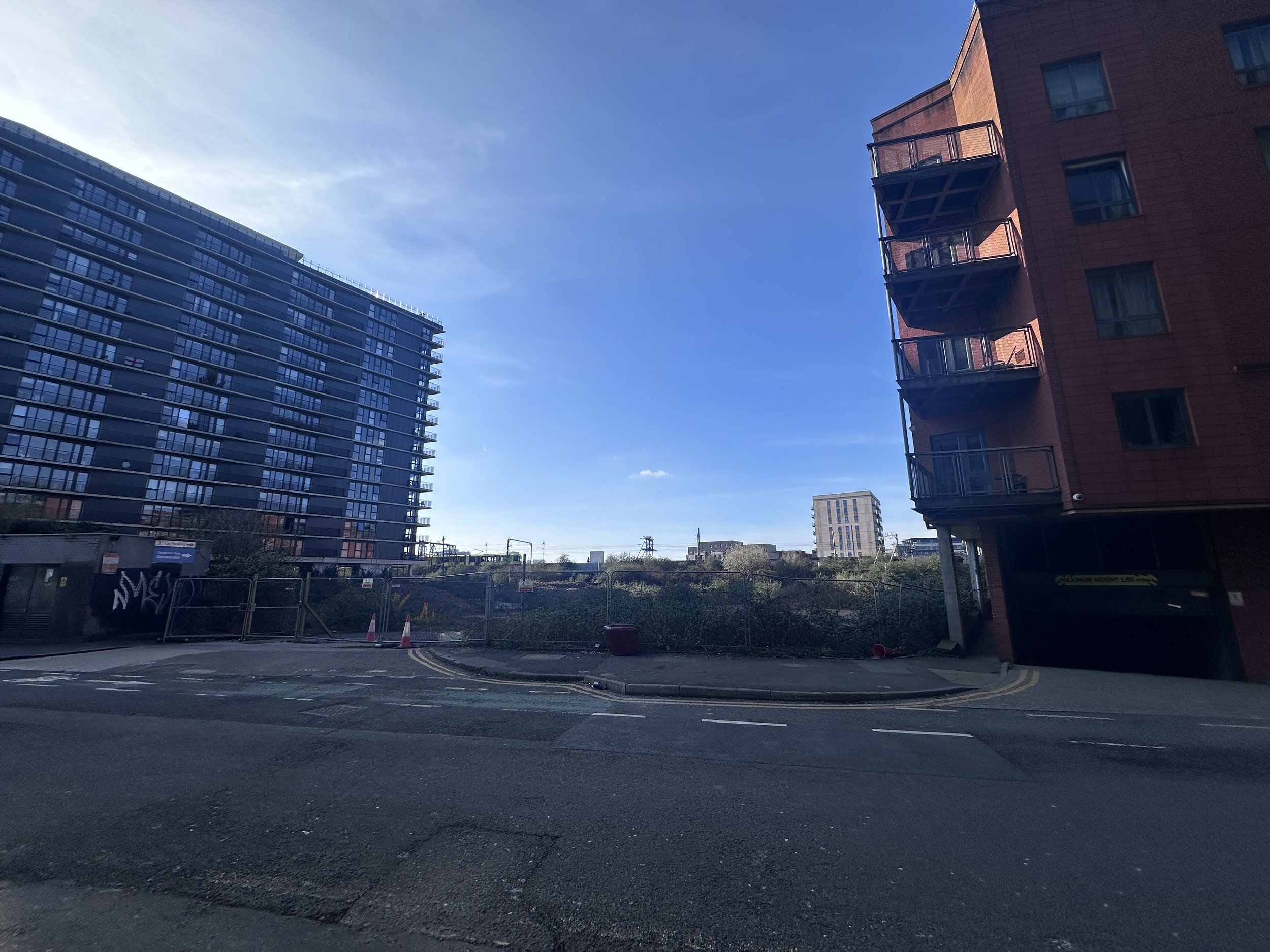 View of a city street with modern buildings, blue sky, and some trees, with a construction site in the foreground.